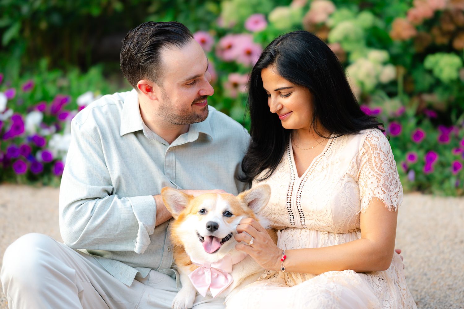 Couple smiling and holding a happy Corgi dog with a pink bow, sitting in a garden with colorful flowers.