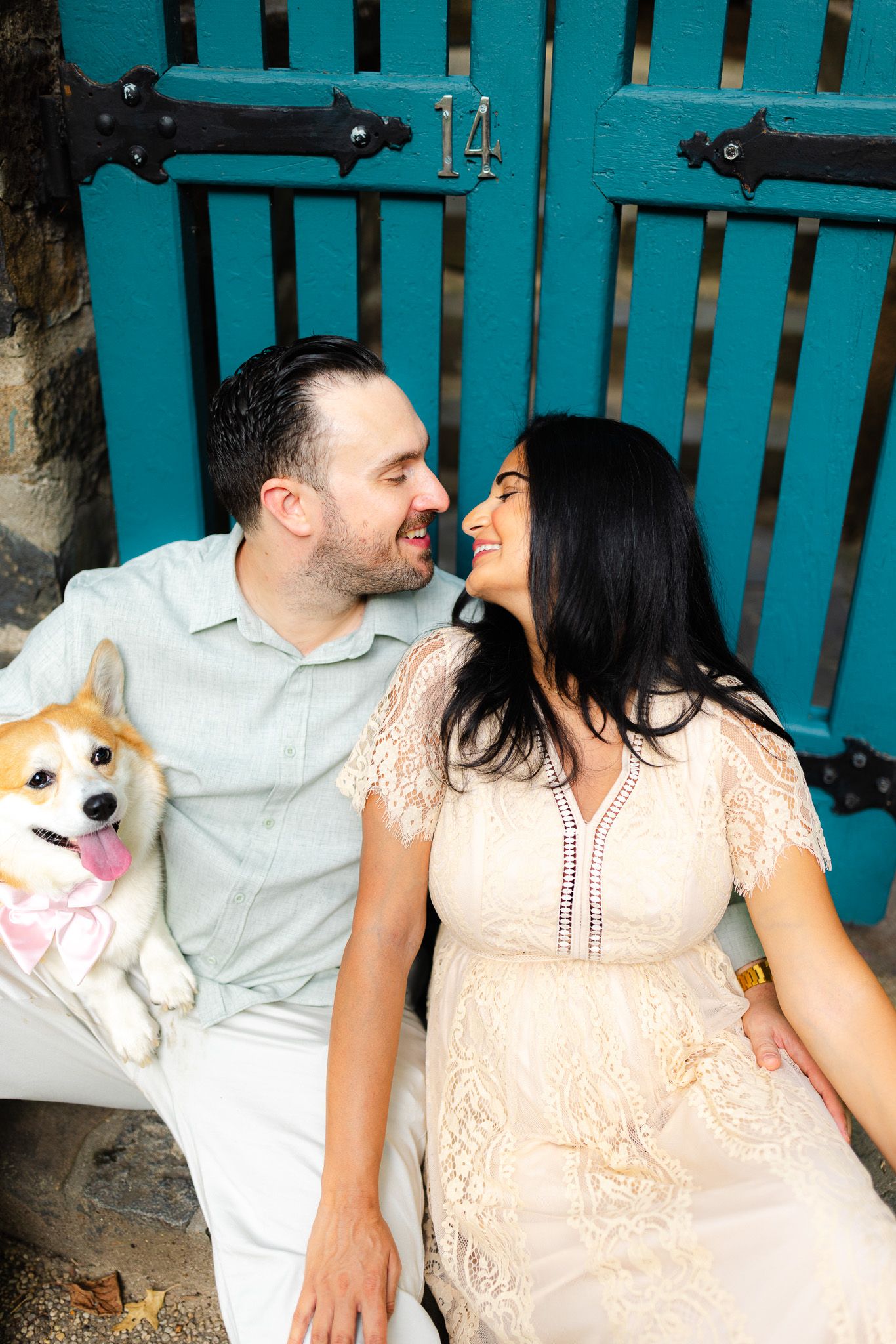Smiling couple sitting together in front of a teal door with a corgi dog, enjoying a cheerful outdoor moment.