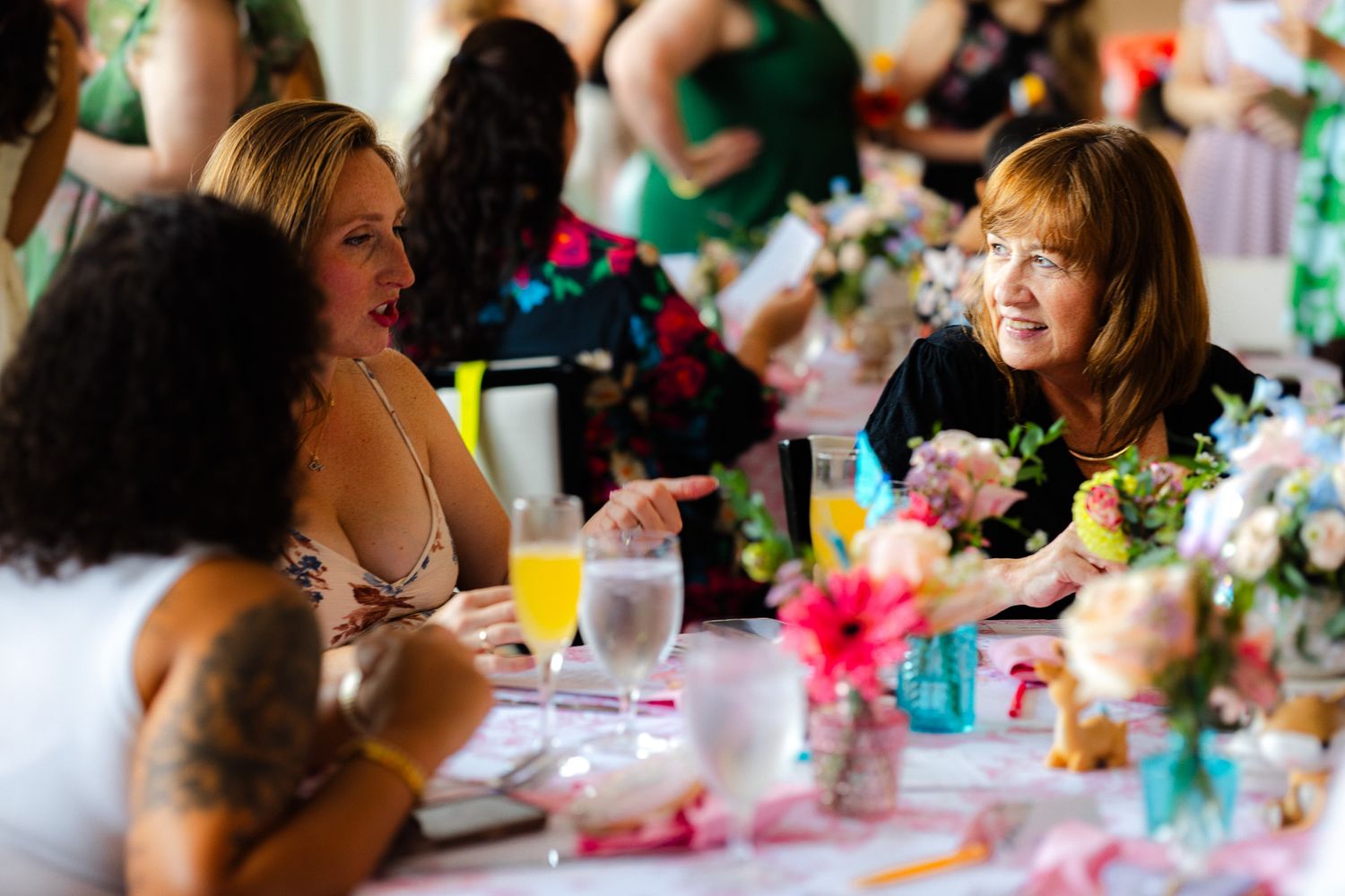 Women in elegant attire laugh and chat at a floral-decorated banquet table with champagne glasses.