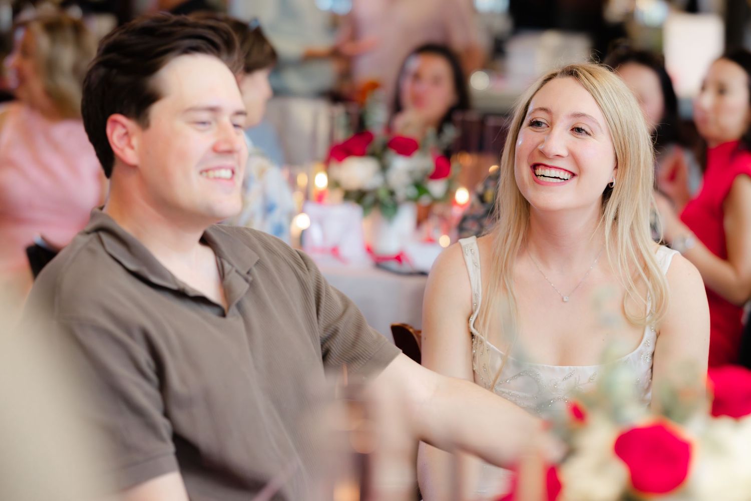 A man and woman laughing together at a festive event with red roses and candles in the background.