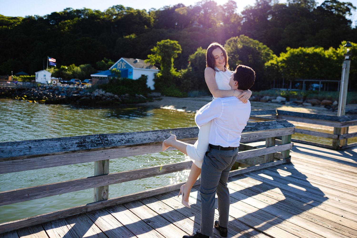 Couple sharing a romantic moment on a sunny waterfront dock, man lifting woman with scenic harbor and trees in background.