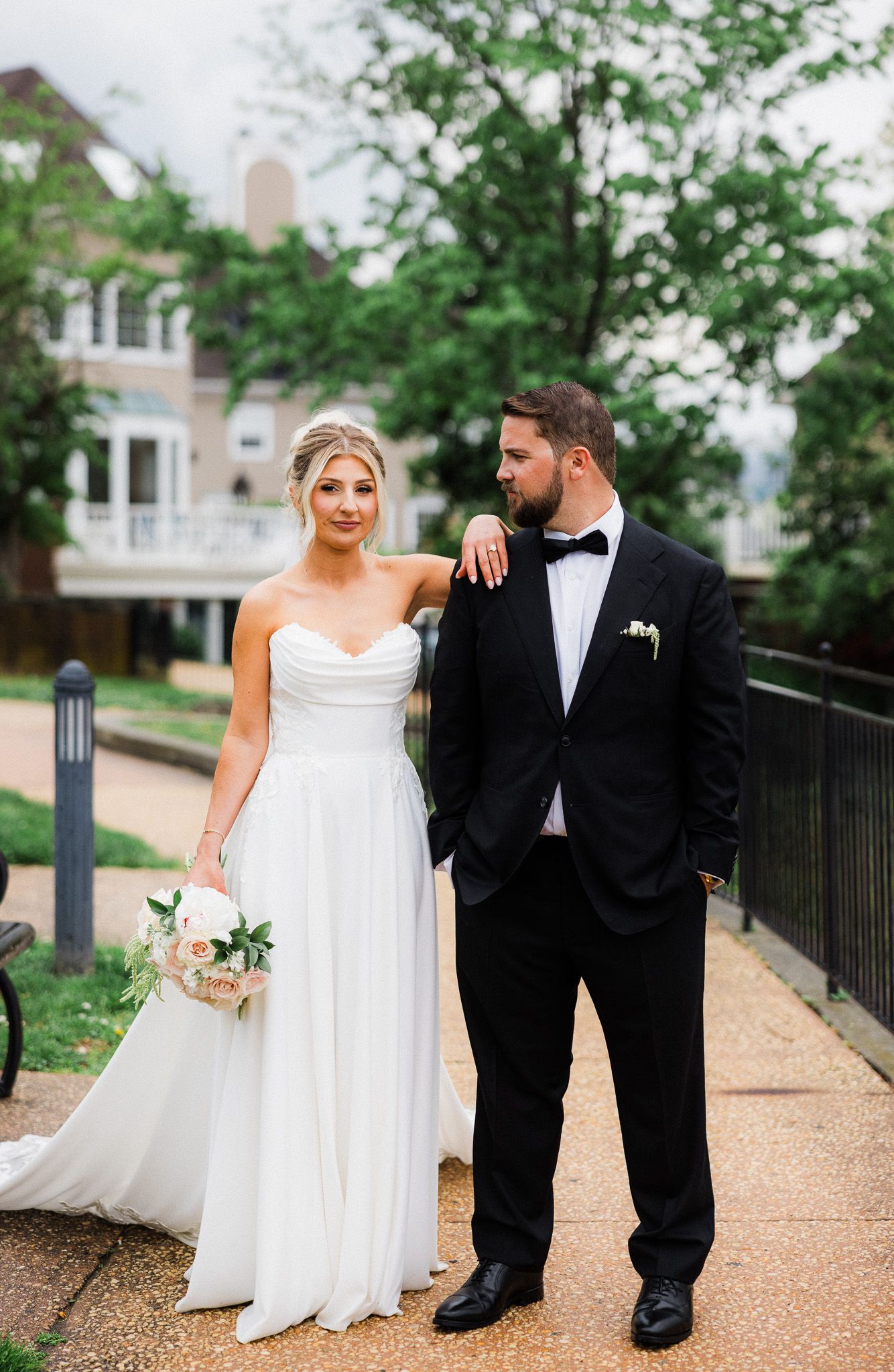 Bride in strapless white gown and groom in black tuxedo pose on outdoor walkway.