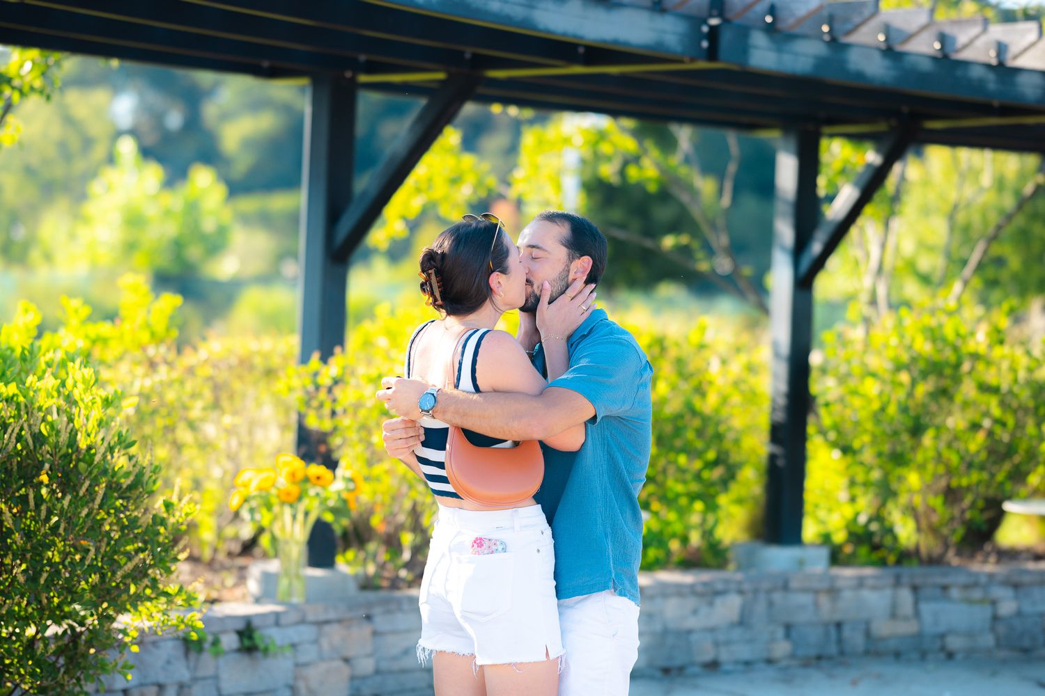 Couple sharing a passionate kiss outdoors under a pergola surrounded by lush green foliage on a sunny day.