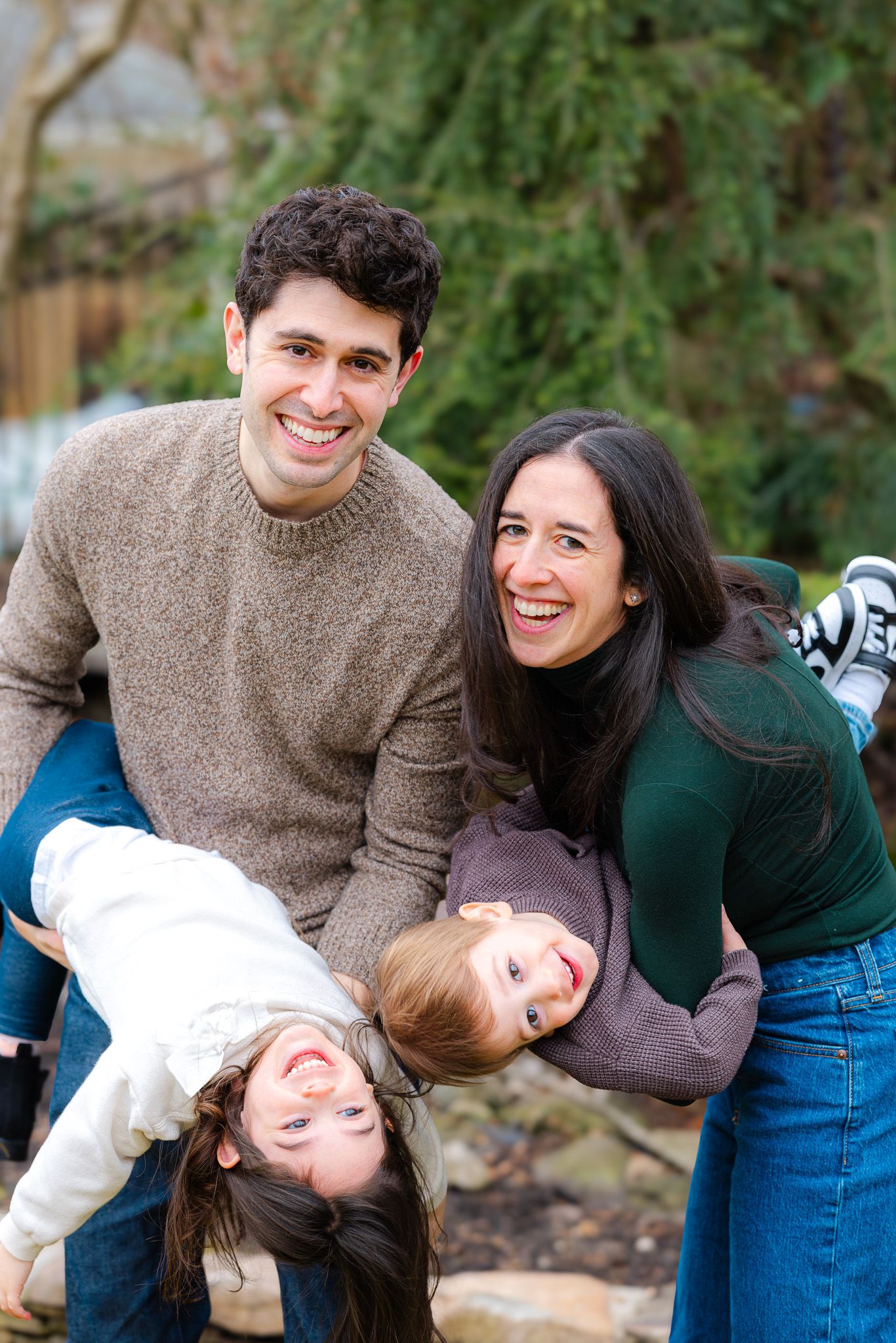 Happy family of four posing outdoors, parents smiling while playfully holding two young children upside down.