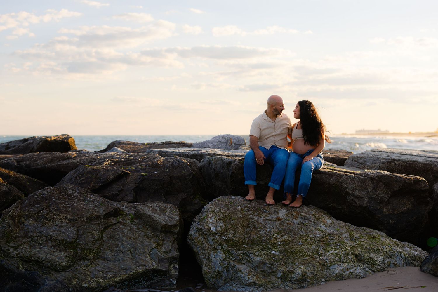 Couple sitting together on rocks by the ocean at sunset, sharing a romantic moment.