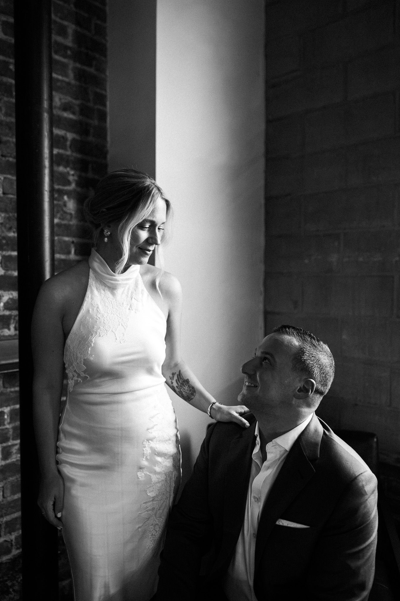 Black and white wedding photo of bride in white gown and groom in suit sharing a romantic moment by a column.
