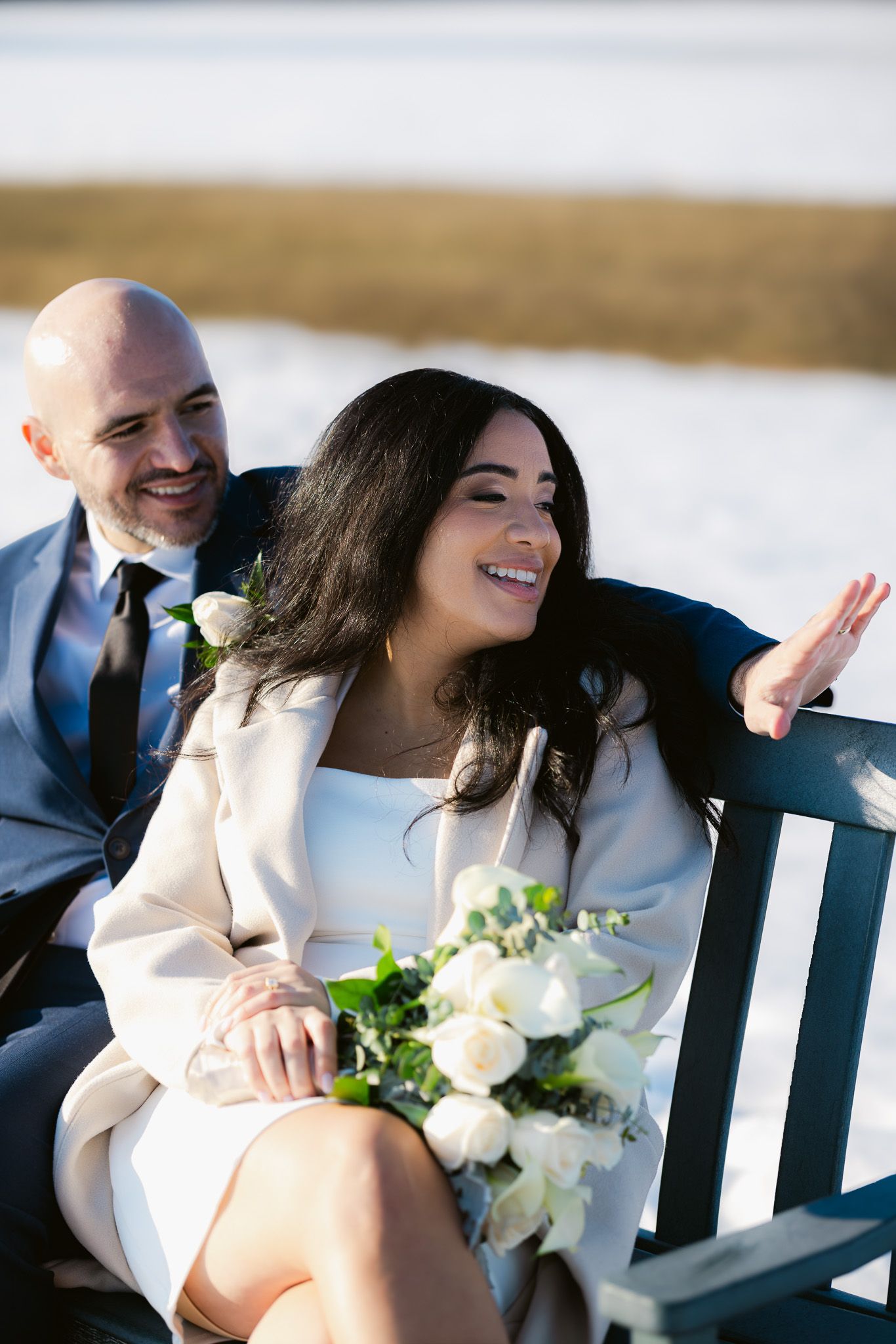 A smiling bride holding white roses sits on a bench with her groom in a suit at a snowy outdoor winter wedding.