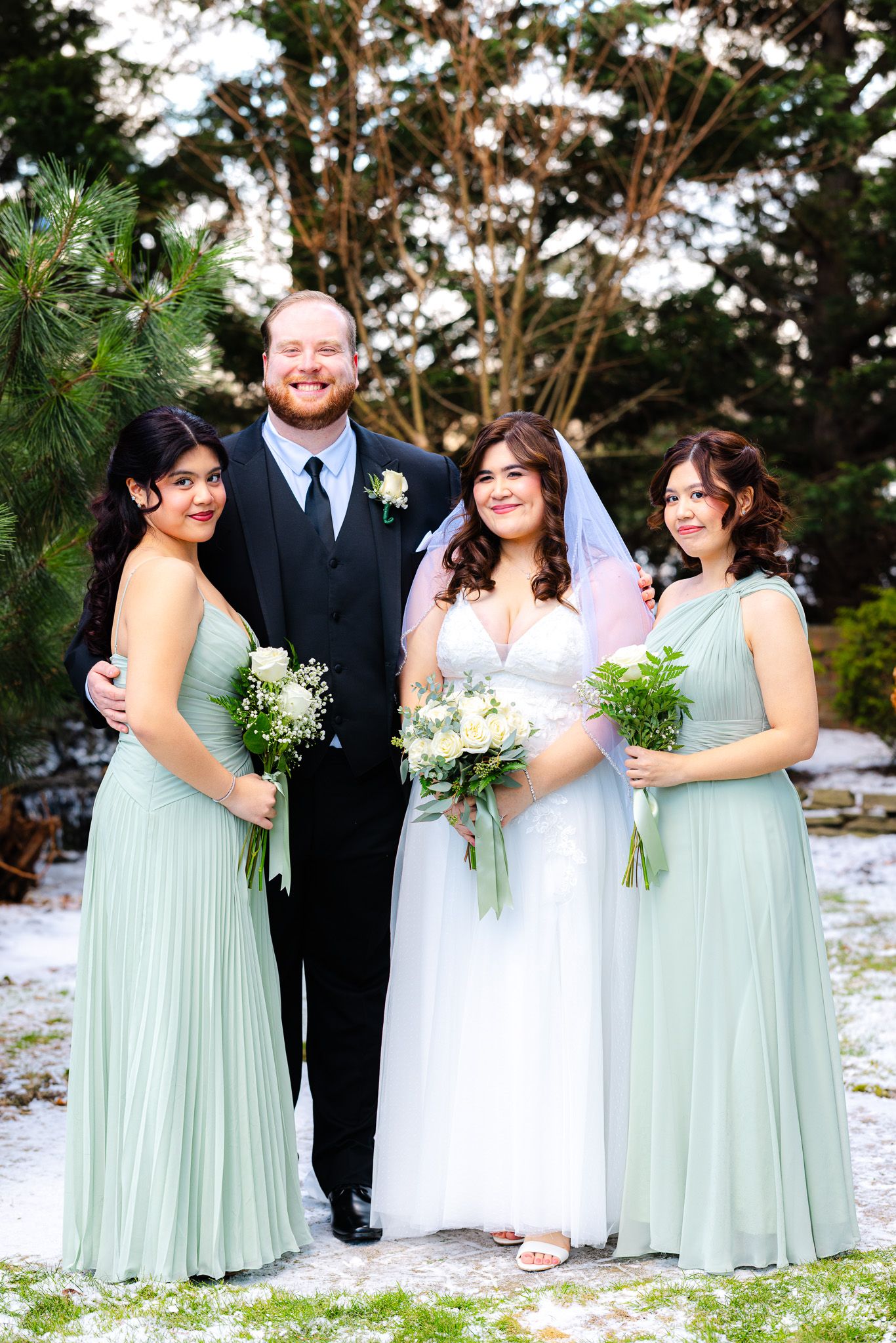 Bride and groom pose with two bridesmaids in sage green gowns holding bouquets outdoors in a snowy winter setting.