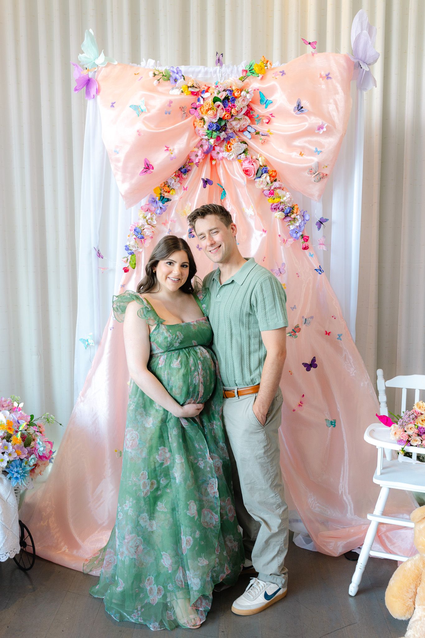 Pregnant couple posing together in front of a pink floral butterfly backdrop at a baby shower celebration.