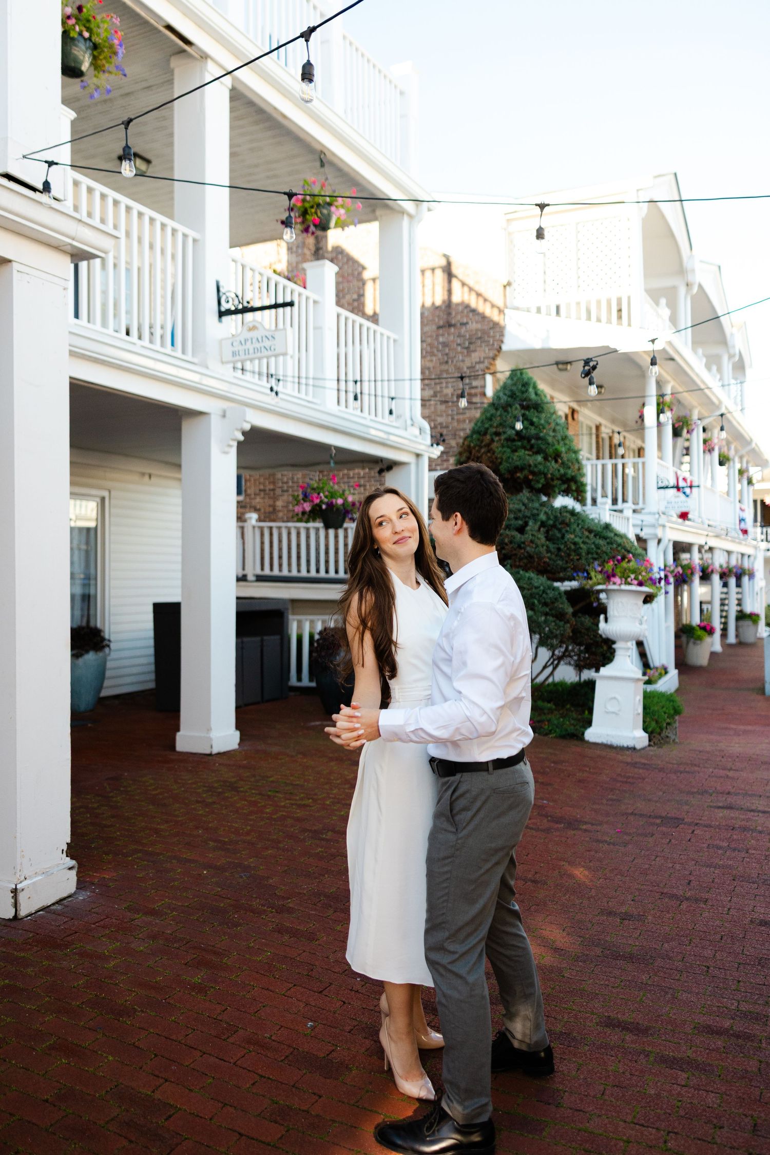 A couple embraces and dances on a brick walkway outside a charming white colonial building with balconies.