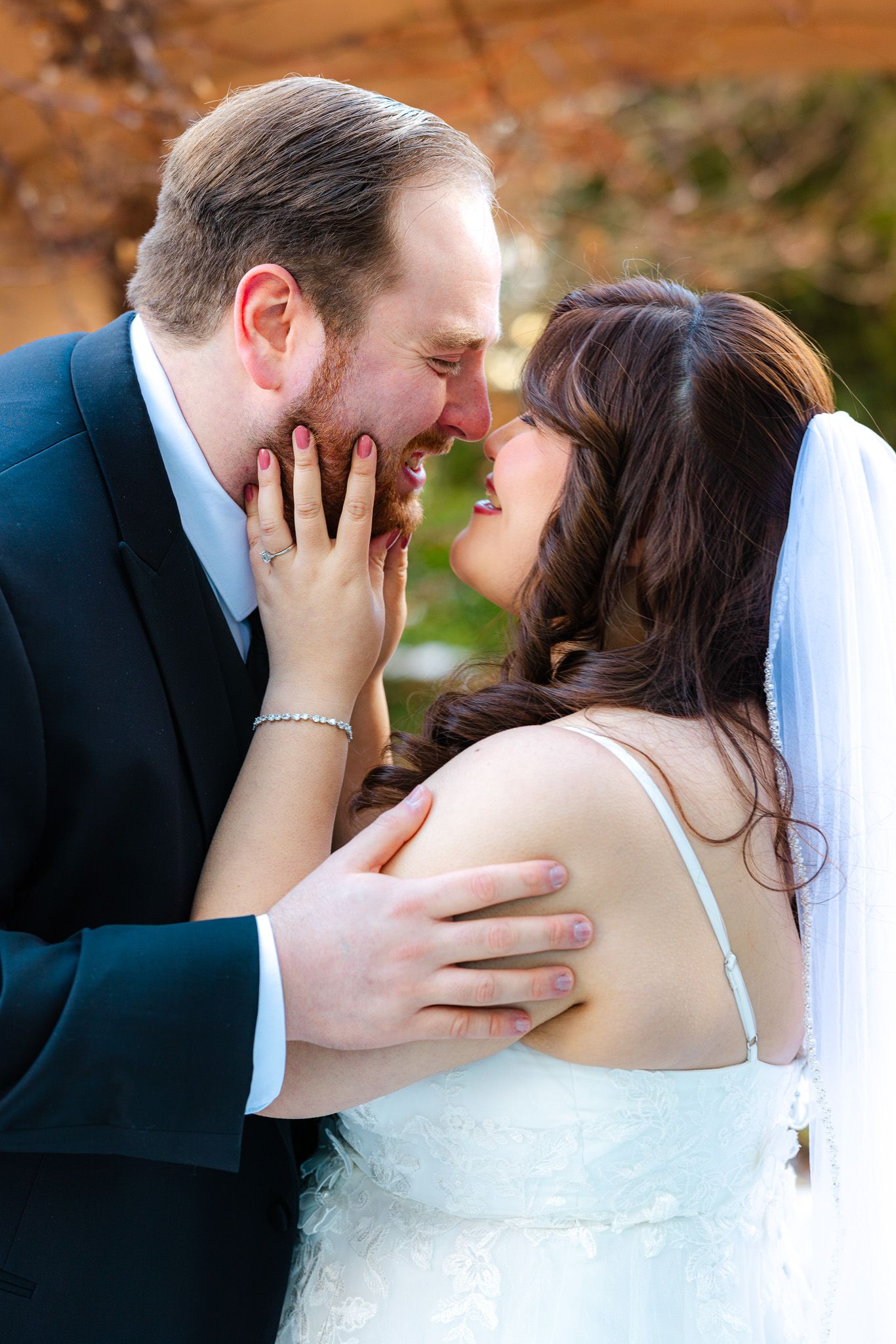 Bride and groom share a romantic almost-kiss moment, with bride in white lace dress and veil outdoors.