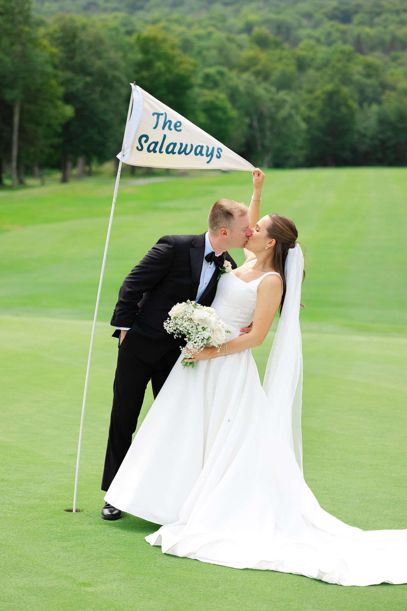 Newlyweds kiss on a golf course holding a custom pennant flag reading The Salaways, bride in white gown with bouquet.