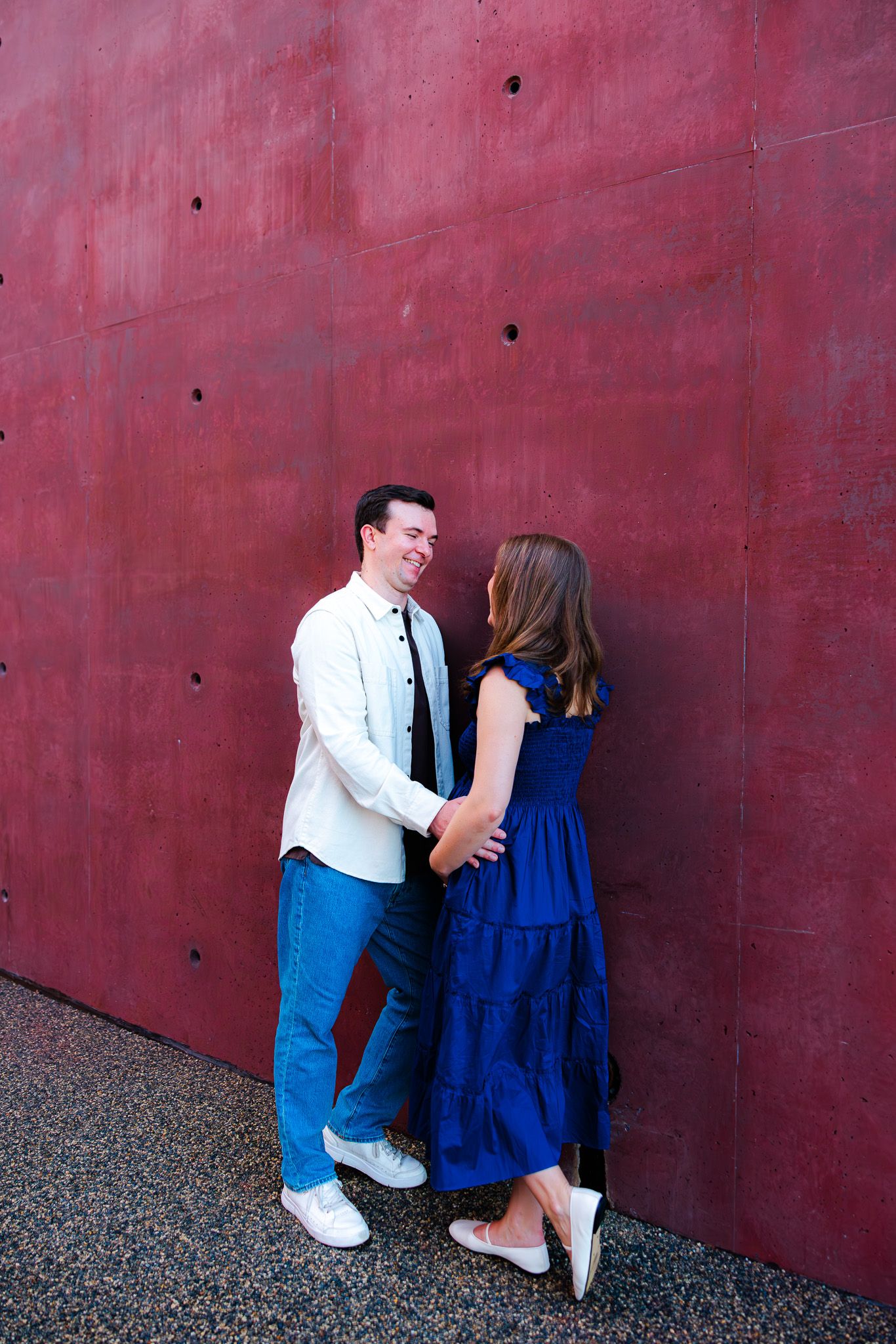 Couple holding hands and smiling against a bold red wall, woman in blue dress, man in white shirt.