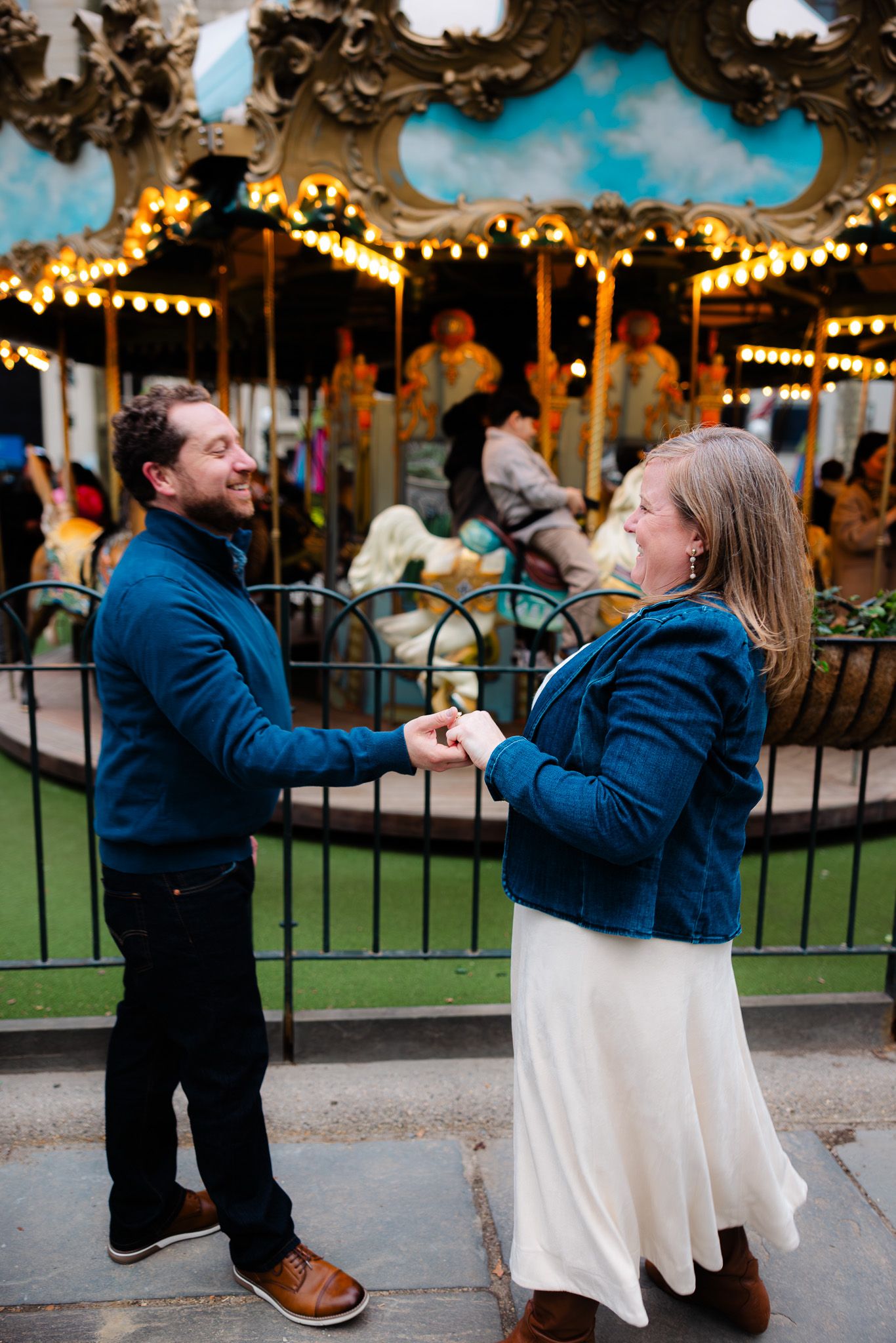 Couple holding hands and smiling in front of a lit carousel in an outdoor setting.