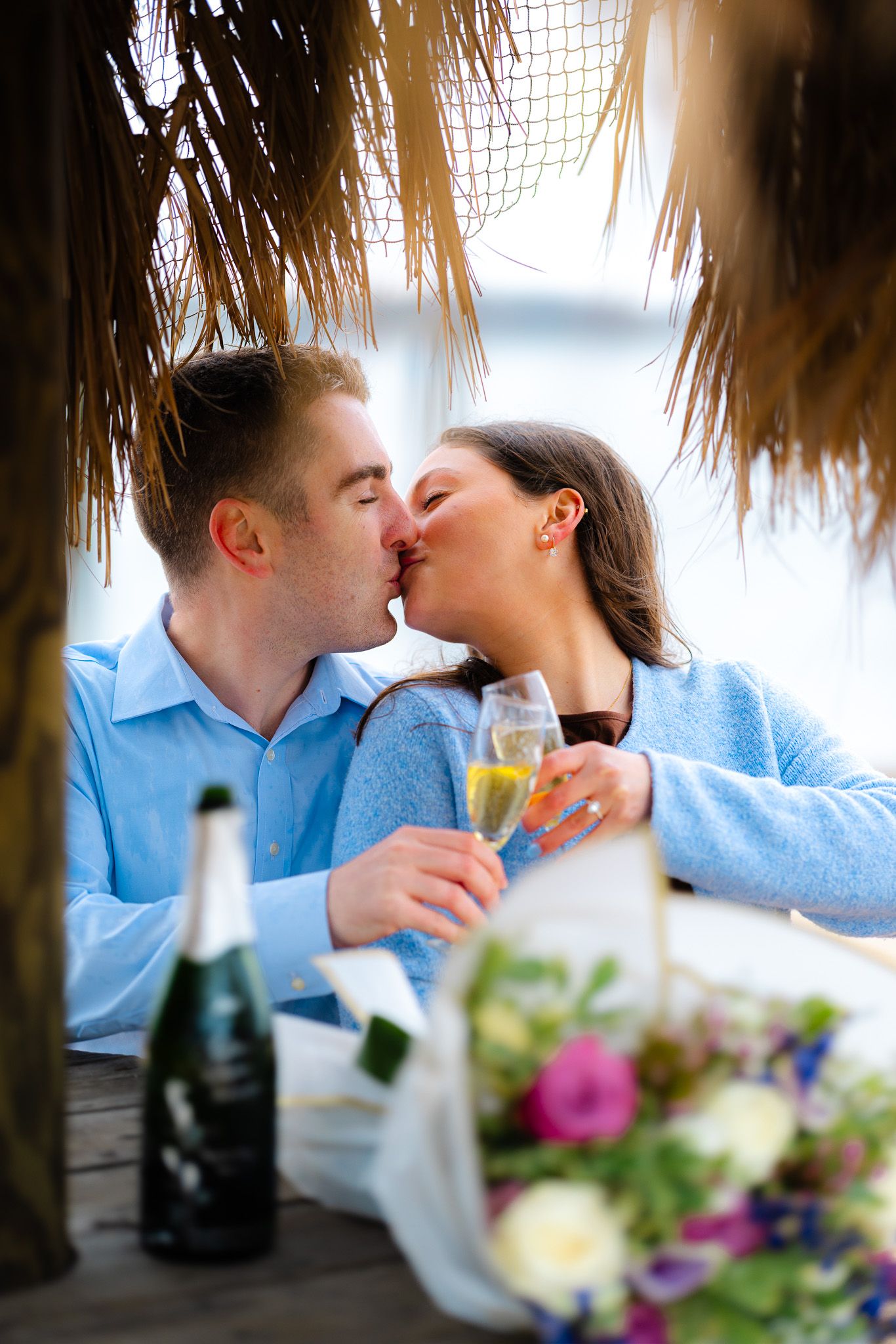 Romantic couple kissing while holding champagne glasses at a beachside table with flowers and a bottle of sparkling wine.