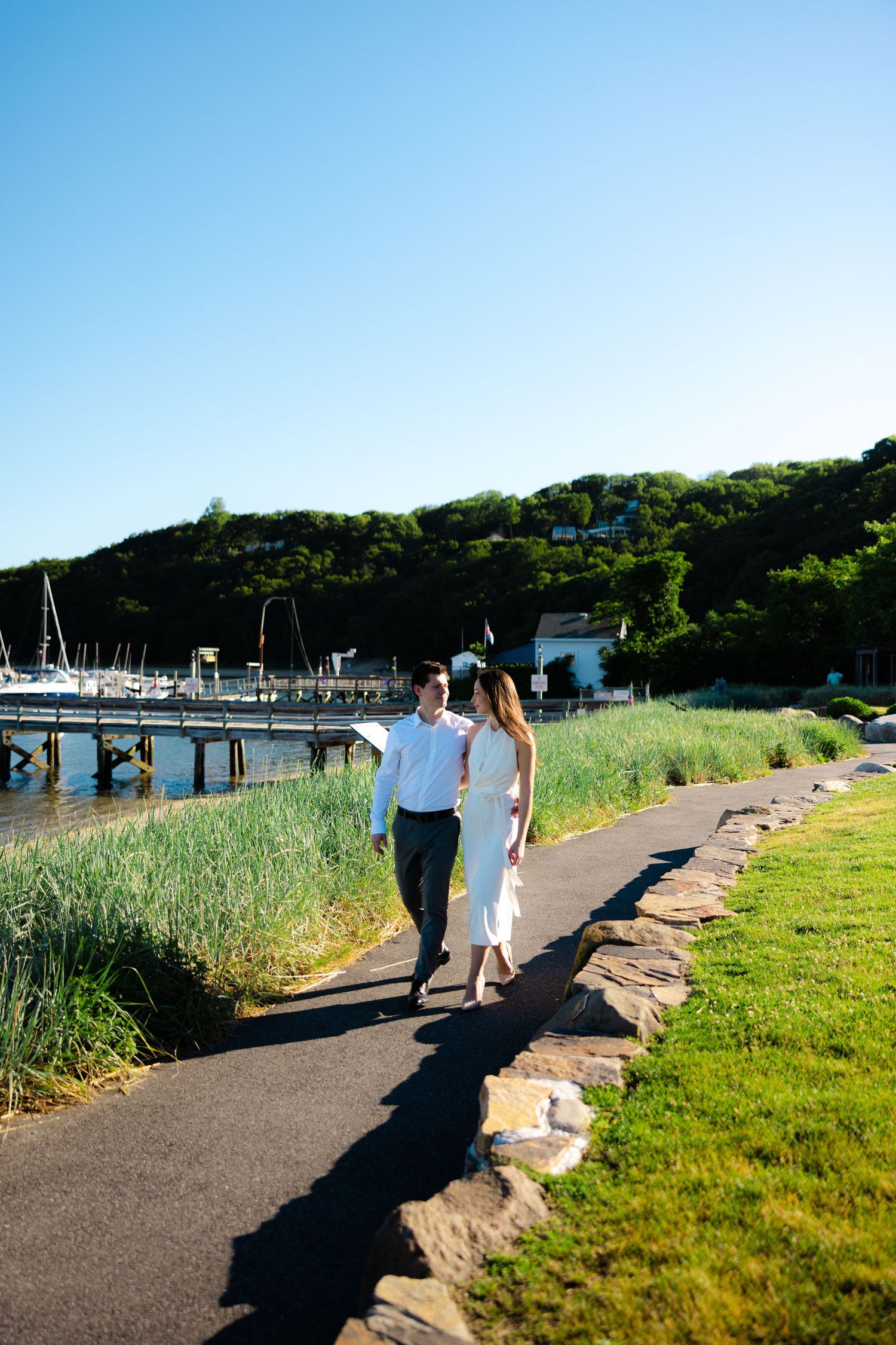 Couple walking hand-in-hand along a waterfront path near a marina with lush green hills and clear blue sky.