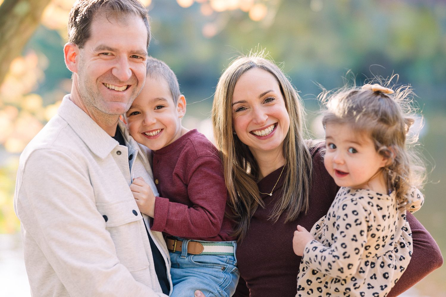 Happy family of four smiling outdoors in autumn, parents holding two young children in a warm, sunlit park setting.