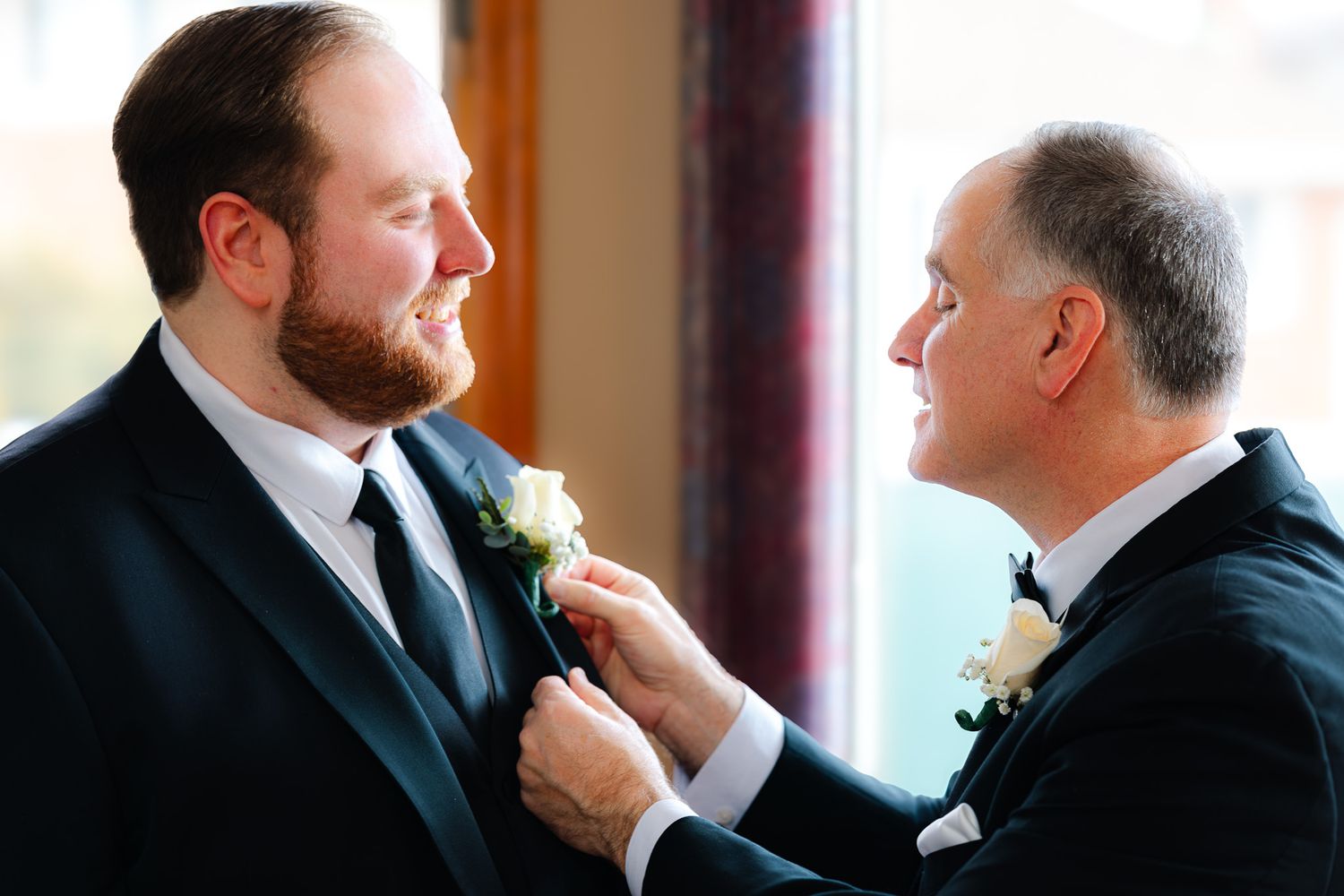 Father pins boutonniere on groom's lapel during wedding preparation, both wearing black suits.