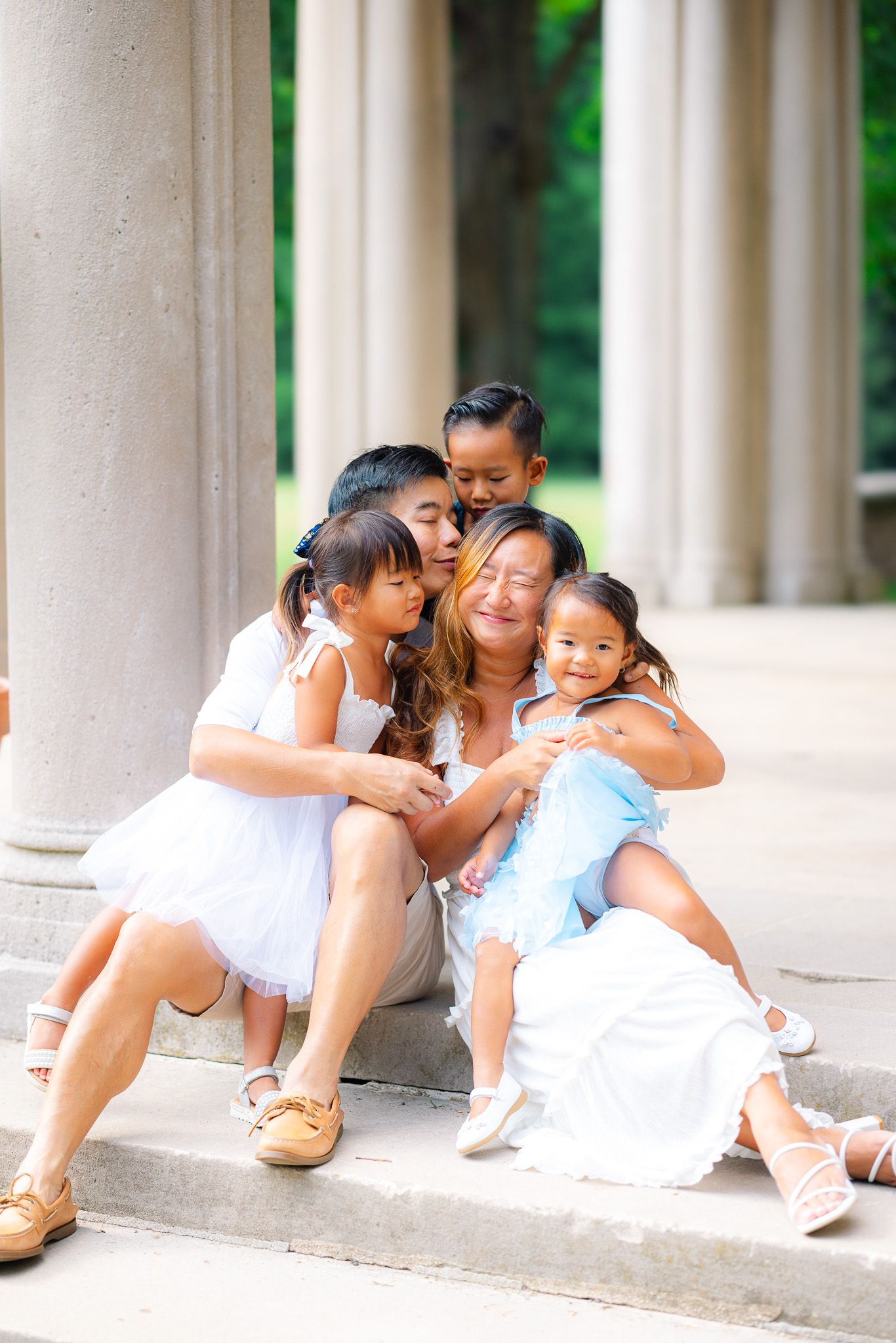 A mother sits on steps with her four children hugging and kissing her, surrounded by columns in a park.
