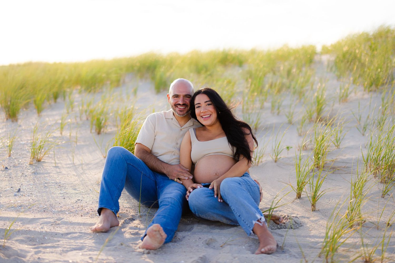 Couple sitting on sandy beach dunes, pregnant woman leaning on man, smiling in warm sunlight.