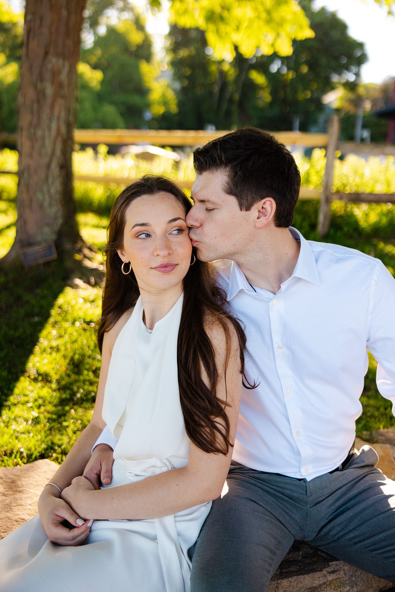 A young man in a white shirt kisses a woman on the cheek while sitting together in a sunny park.