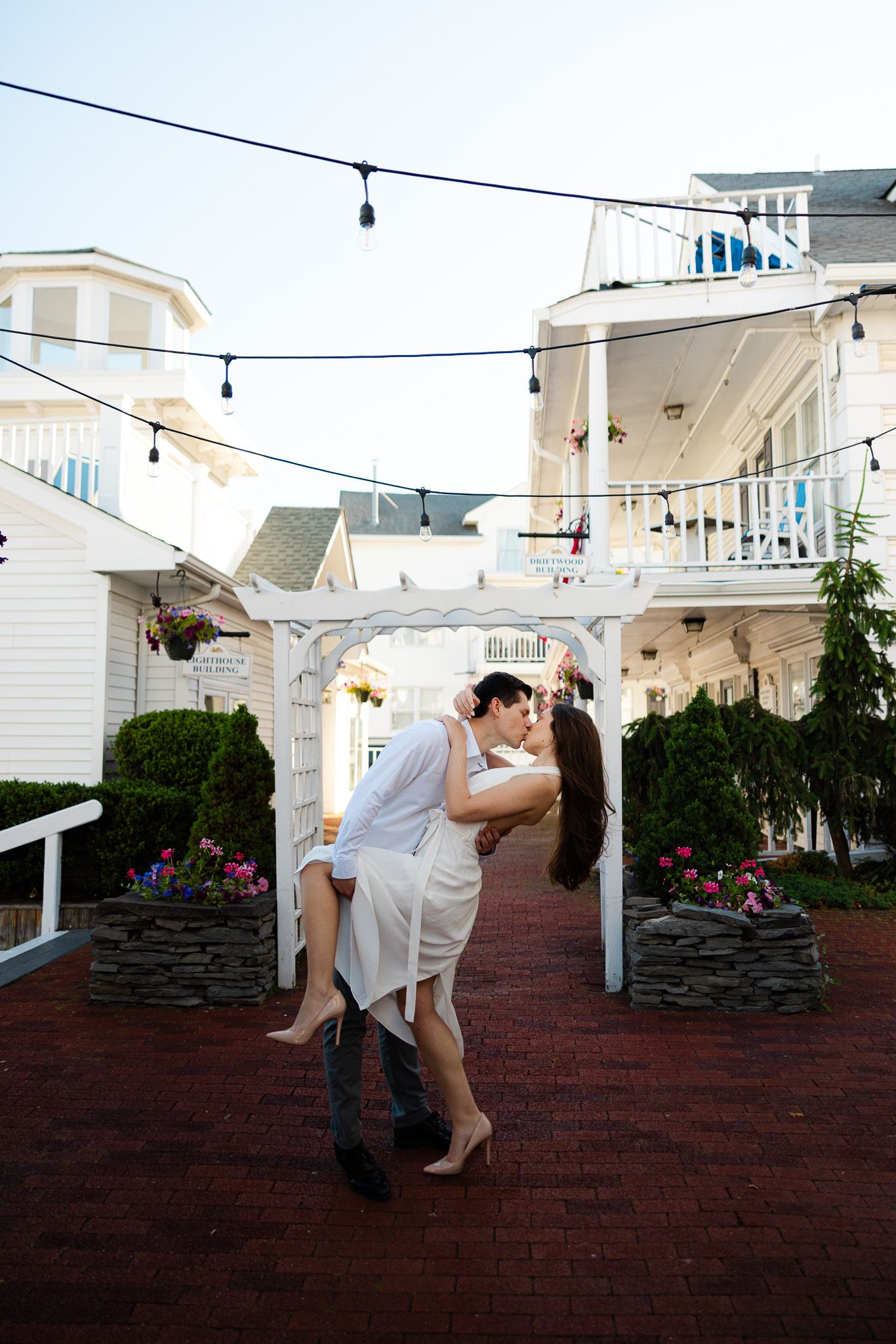 Couple sharing a romantic dip kiss under string lights in a charming courtyard with white buildings and flower gardens.