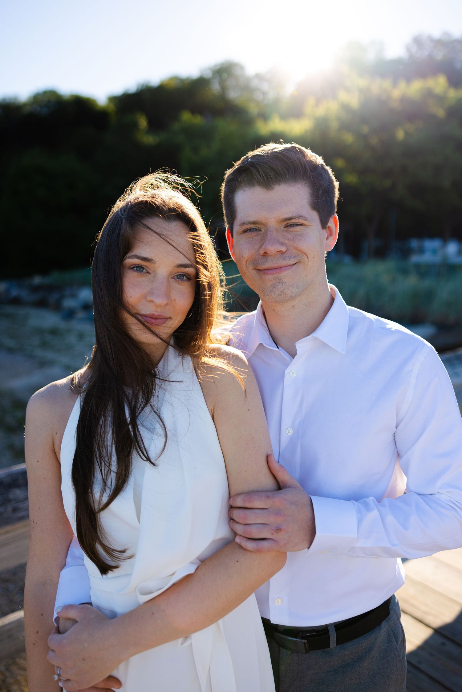 A couple embraces outdoors at golden hour, woman in white dress with long dark hair, man in light blue shirt.