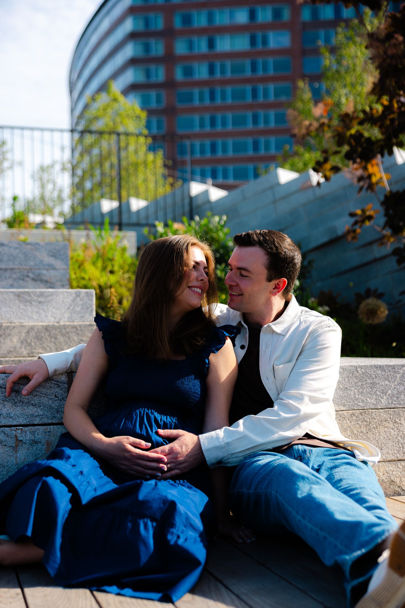 Pregnant couple sitting together on stone steps, smiling at each other with a modern building in the background.