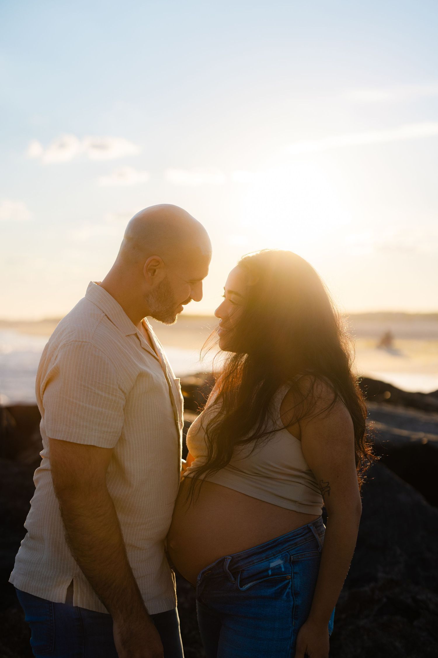Couple sharing intimate moment at sunset beach, pregnant woman cradling baby bump with ocean waves glowing behind them.