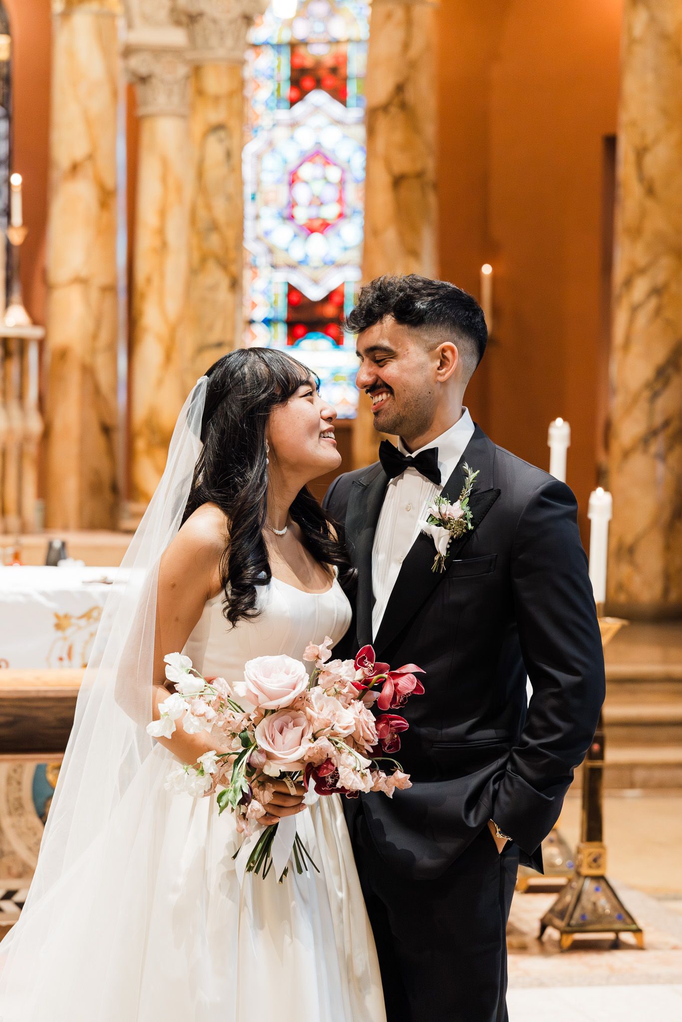 Bride and groom share a joyful moment inside a church, bride holding pink and red bouquet, groom in black tuxedo.