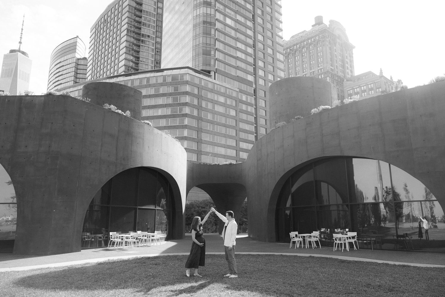 Couple dancing outdoors near modern arched architecture with city skyscrapers in black and white photo.