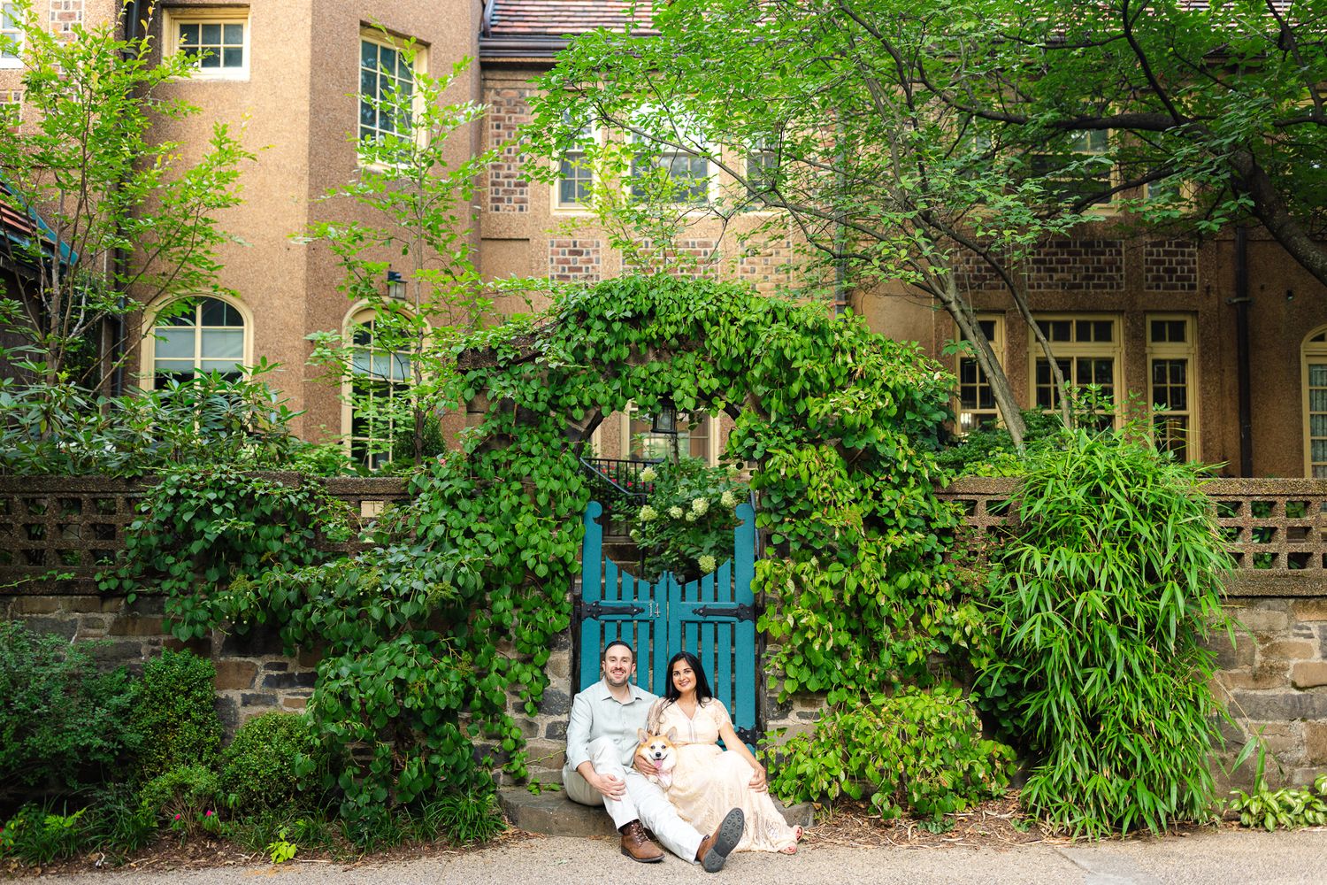 Couple sitting together in front of ivy-covered stone building with blue garden gate surrounded by lush greenery.