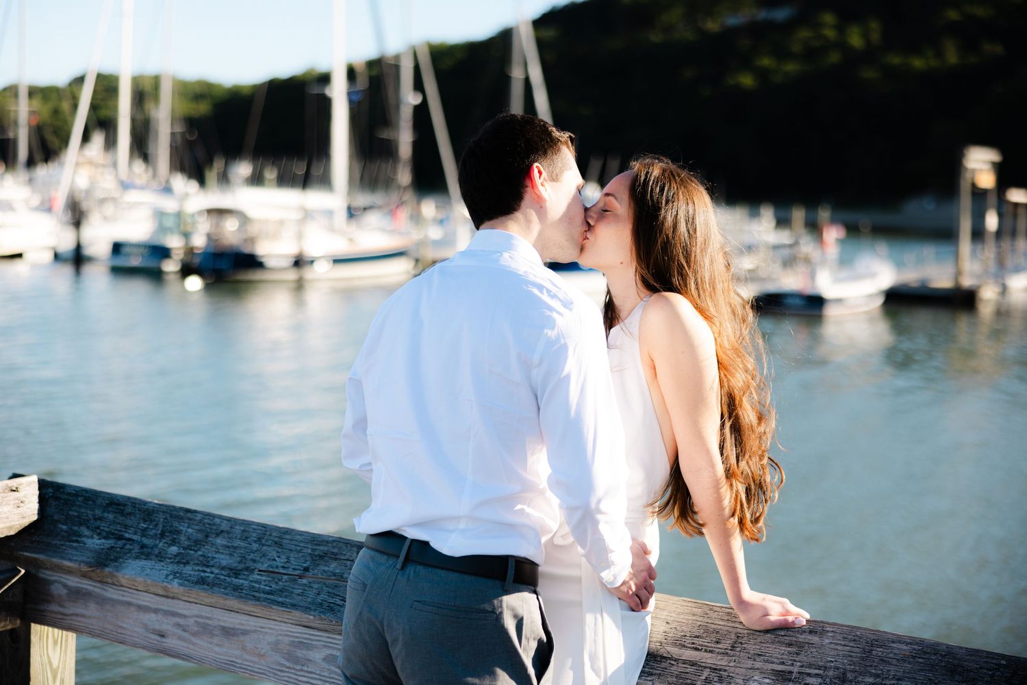 Couple sharing a romantic kiss on a waterfront dock with sailboats and marina in the sunny background.