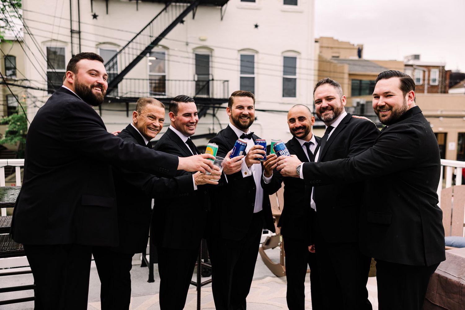 Seven groomsmen in black suits toasting with drinks outdoors on a rooftop terrace before a wedding celebration.