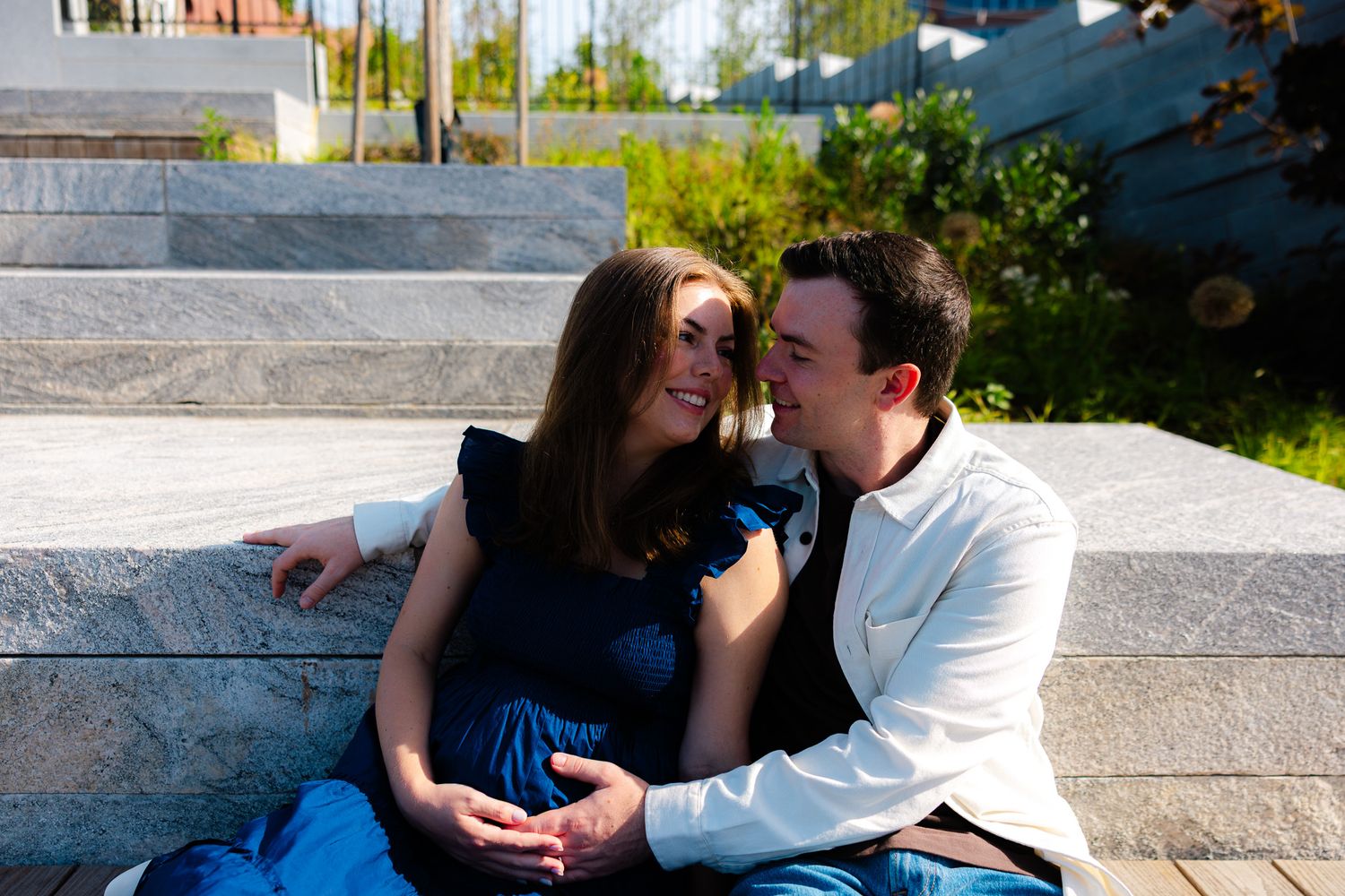 A happy couple sits on outdoor stone steps, sharing a tender moment, with the woman in a blue dress cradling her baby bump.