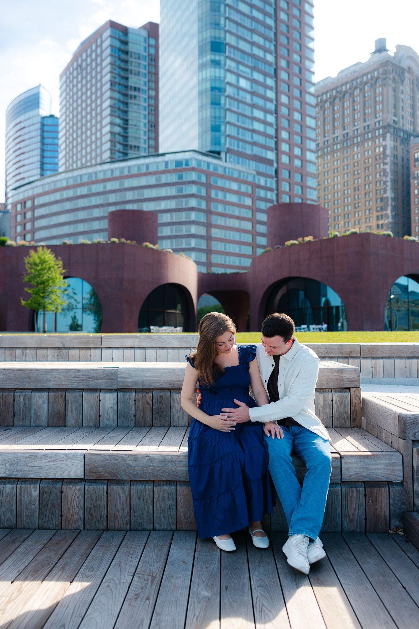 Pregnant couple sitting on wooden steps with city skyscrapers backdrop, woman in blue dress, man in white shirt.