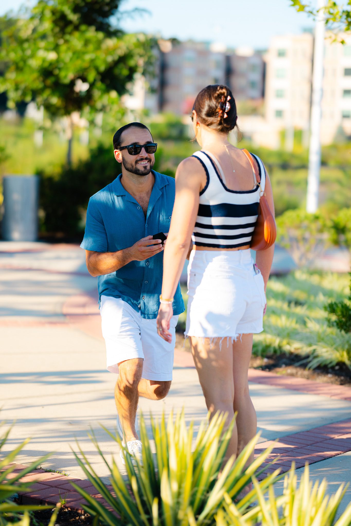 Couple walking together in a sunny park, man in blue shirt and white shorts smiling at woman in striped top.