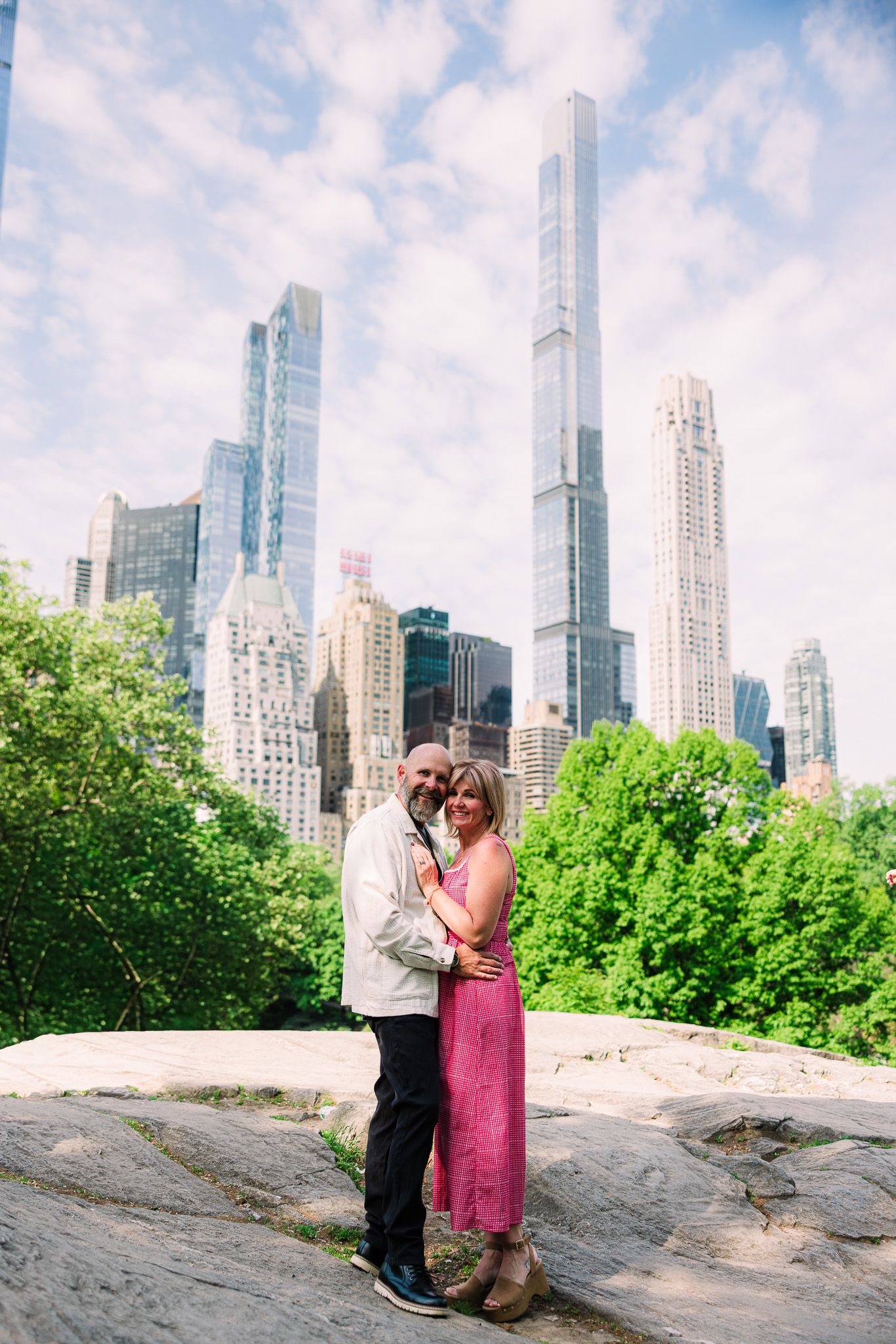 Couple embracing on a rocky outcrop in Central Park with the Manhattan skyline visible behind them.