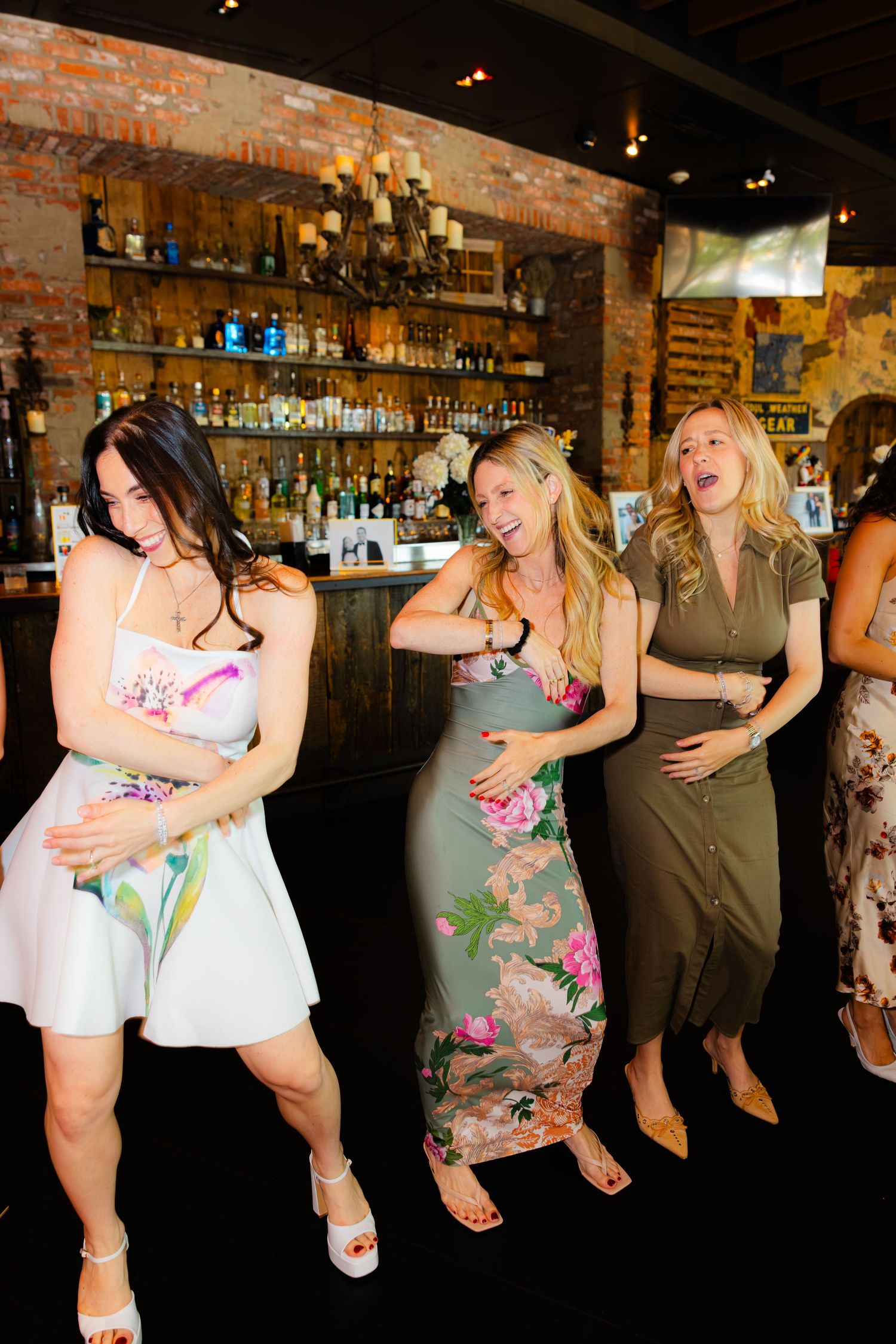 Three women dancing and laughing together at a bar venue with brick walls and a chandelier.