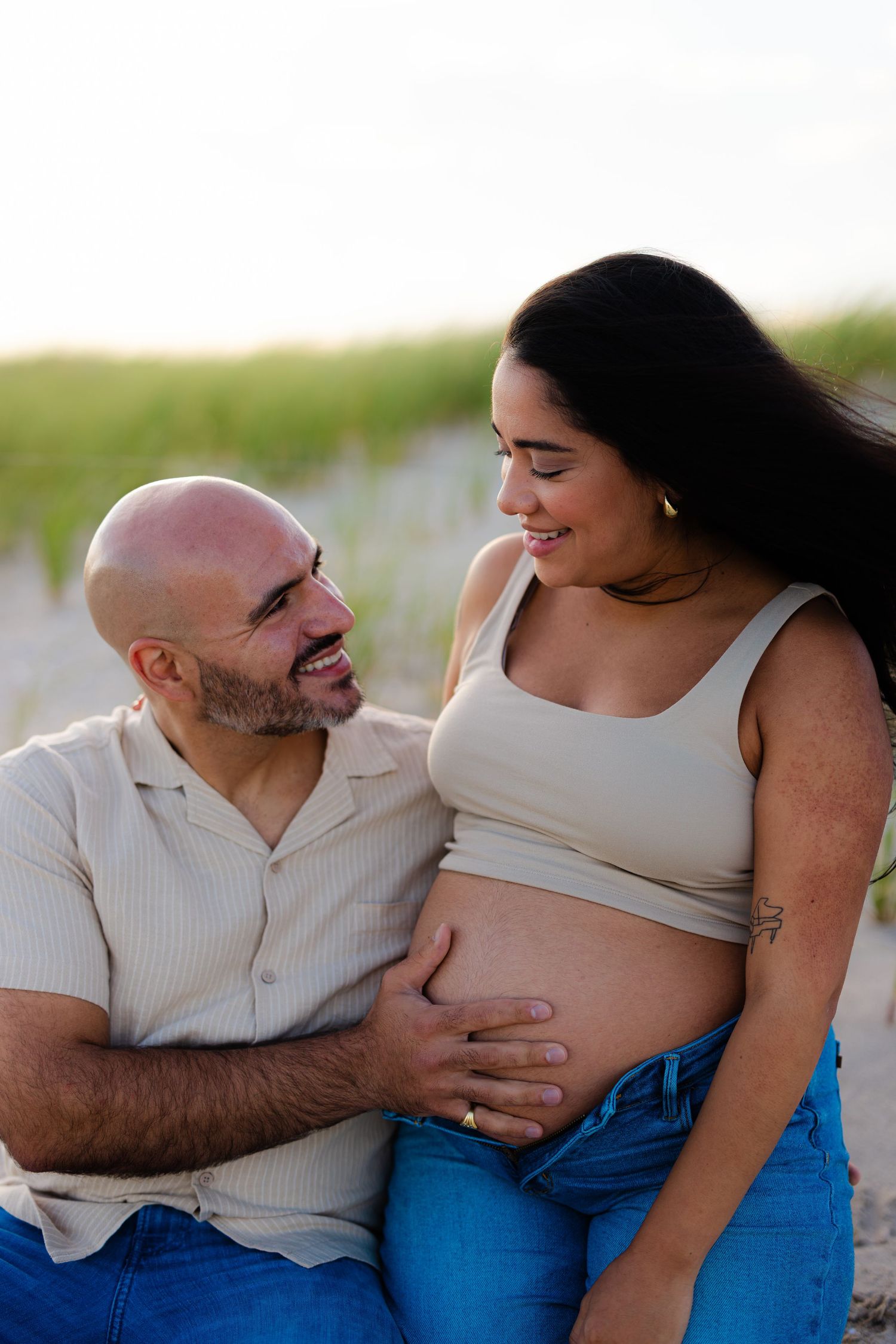 Smiling expecting couple sitting on beach sand, man gazing at pregnant woman as she holds her bare baby bump.