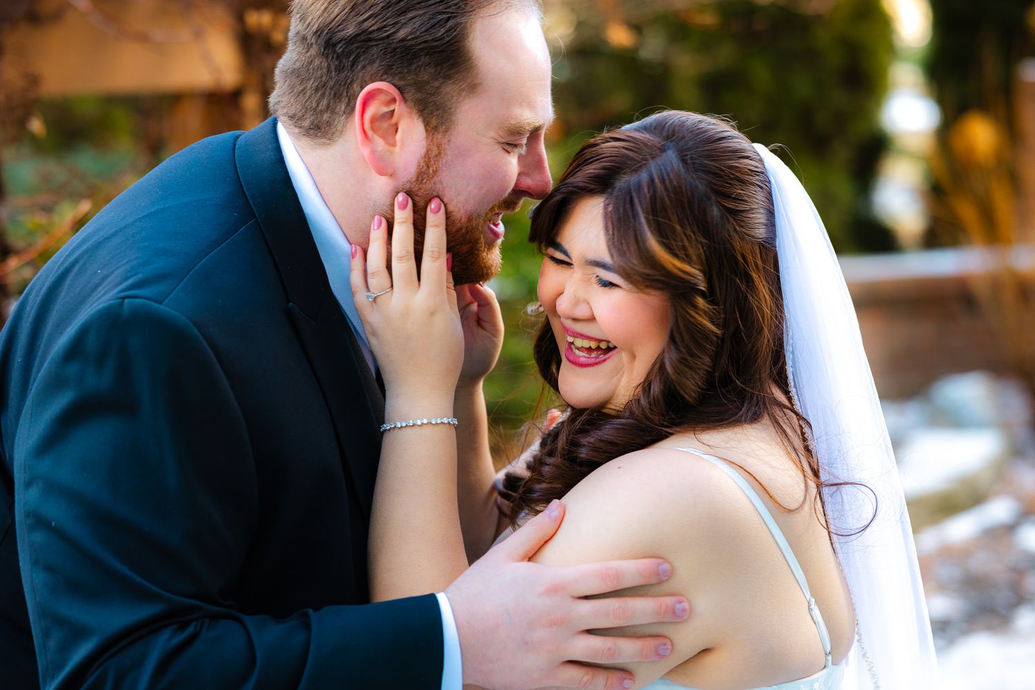 Joyful bride and groom sharing a tender moment, laughing together outdoors in wedding attire.
