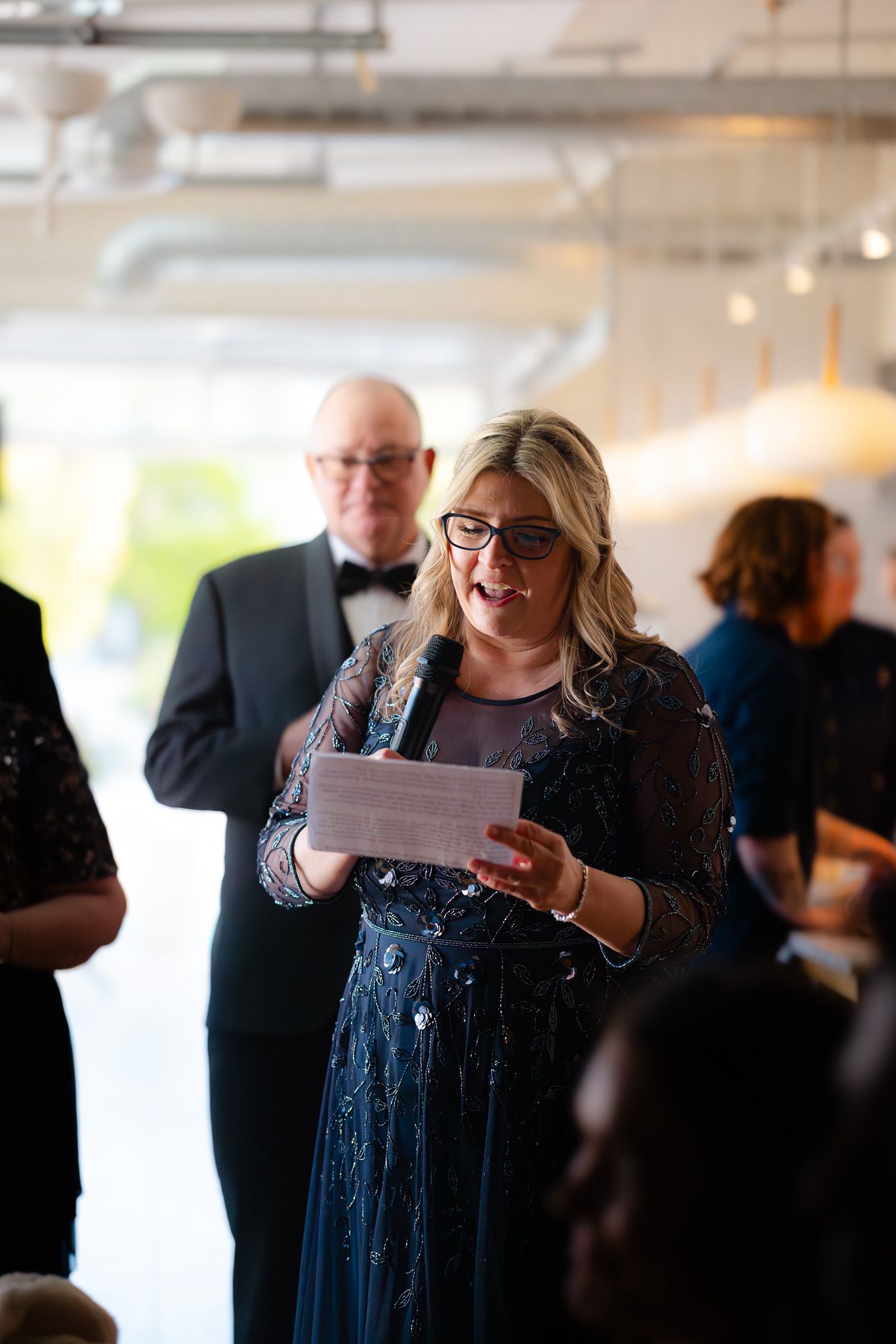 Woman in navy sequin dress reads speech at formal event, man in tuxedo stands behind her.