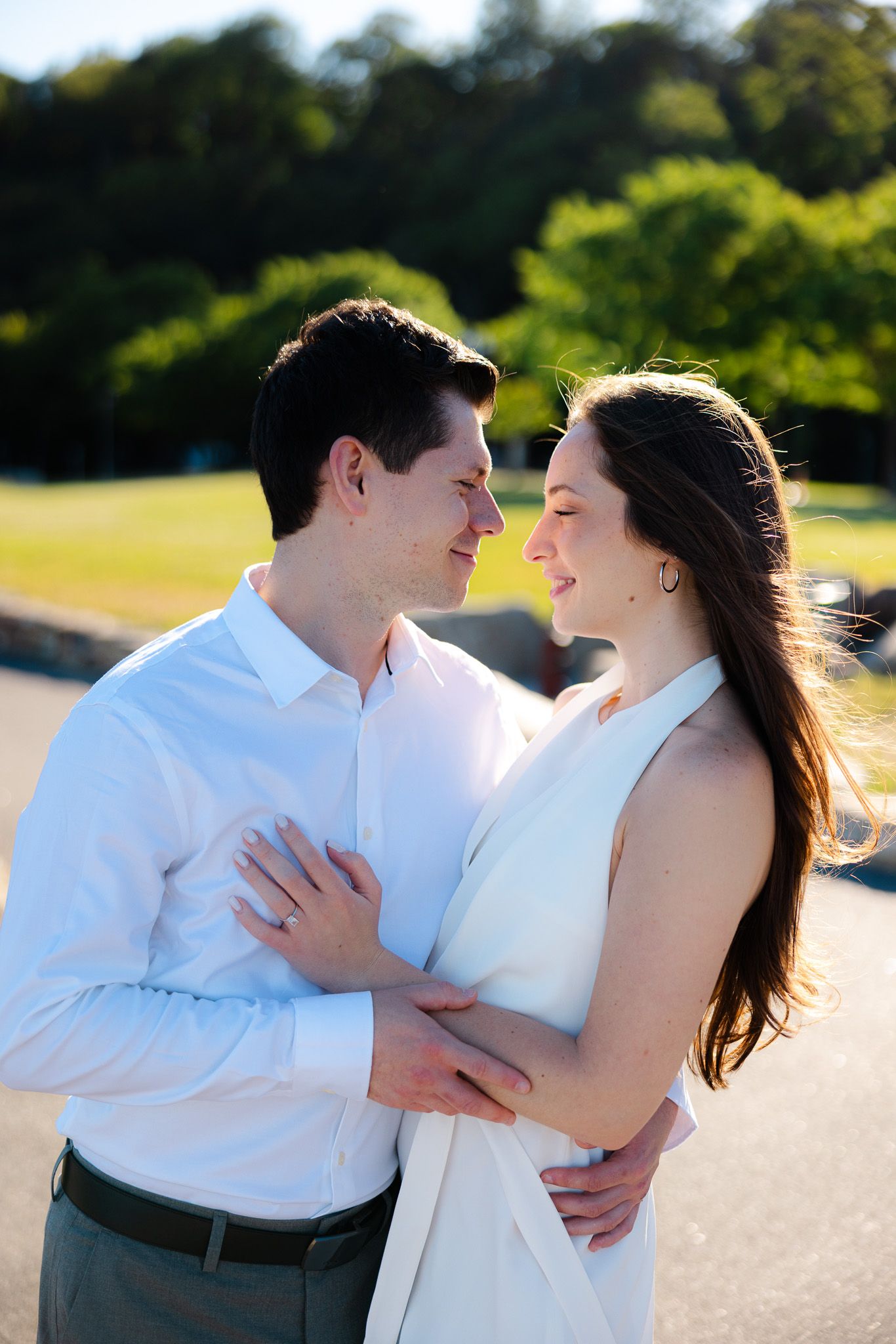 A couple in white attire embraces and gazes lovingly at each other outdoors with green trees in the background.