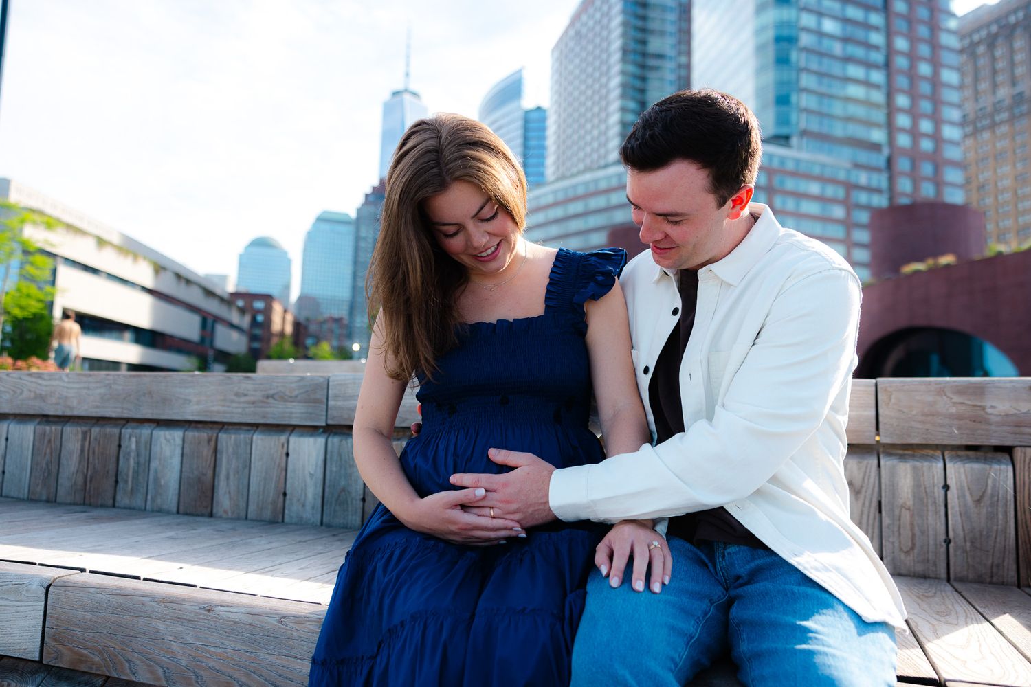 Pregnant couple sitting outdoors with city skyline, woman in blue dress, man in white jacket touching her baby bump.