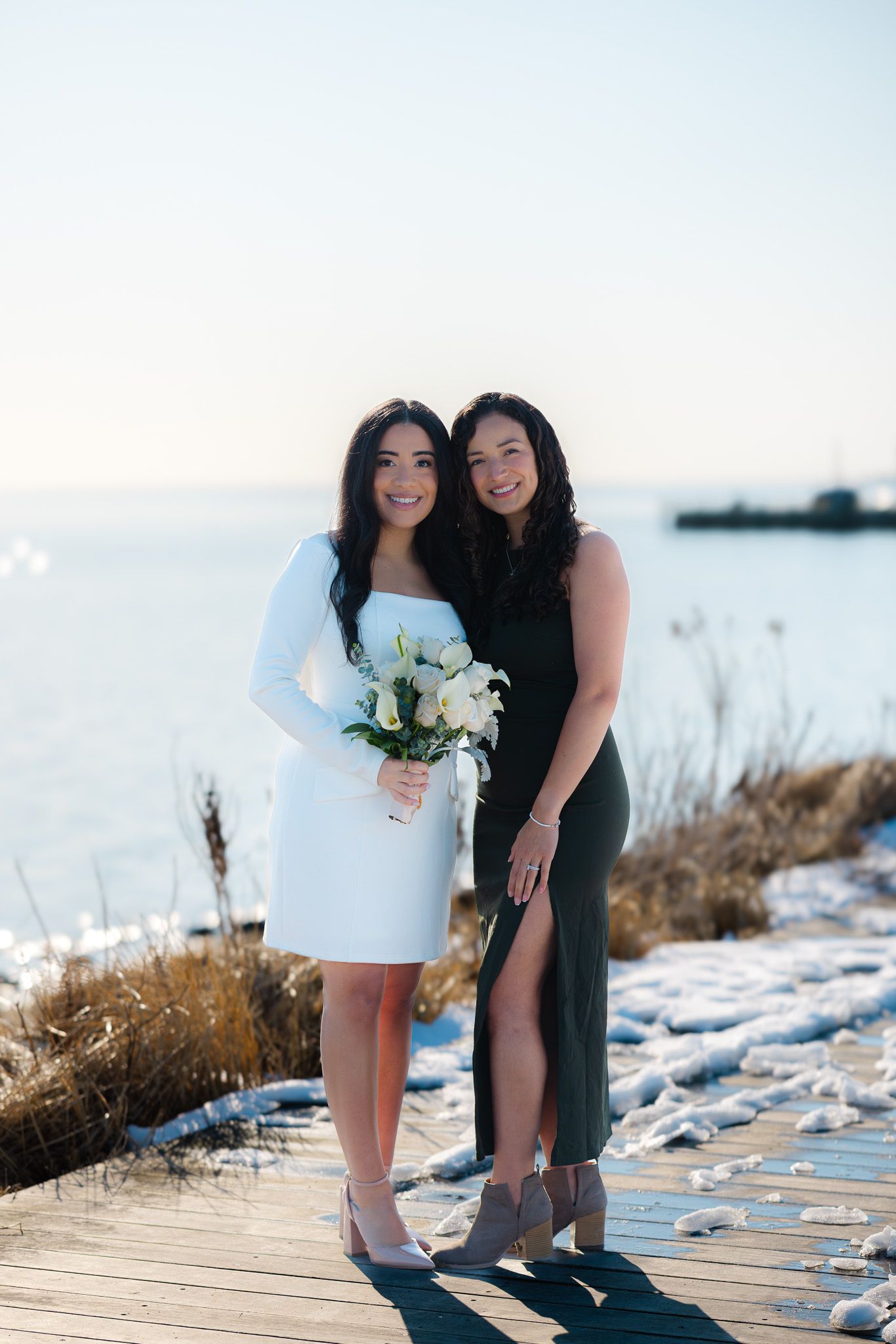 Two women in formal dresses pose on a snowy waterfront boardwalk, one holding a white bouquet.