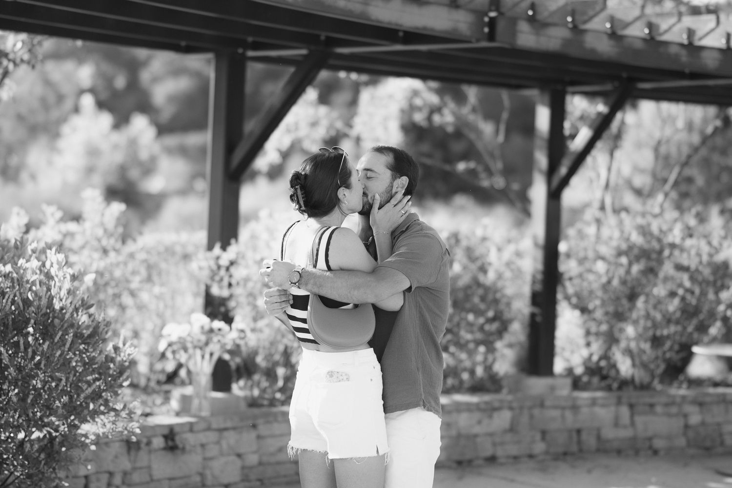 Black and white photo of a couple sharing a romantic kiss outdoors under a pergola surrounded by greenery.