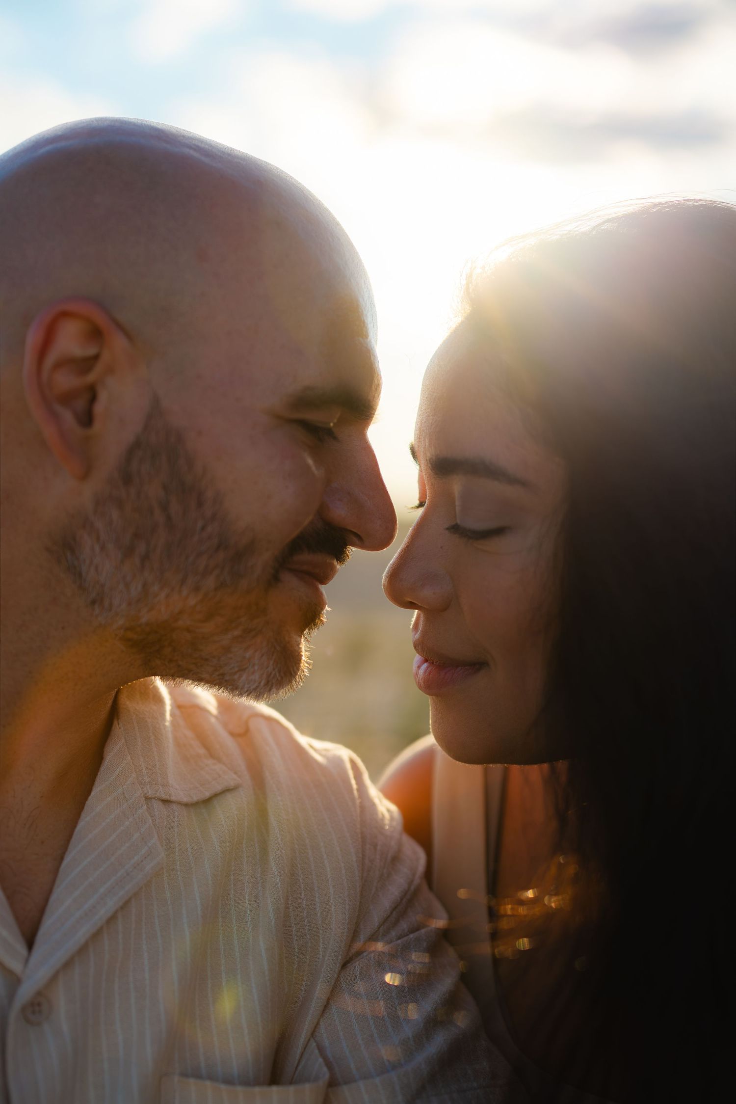 A couple shares an intimate moment at golden hour, foreheads nearly touching, bathed in warm sunset backlight.