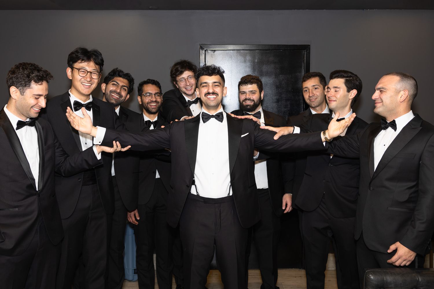 A groom in a tuxedo poses with arms outstretched surrounded by smiling groomsmen in black tie attire indoors.