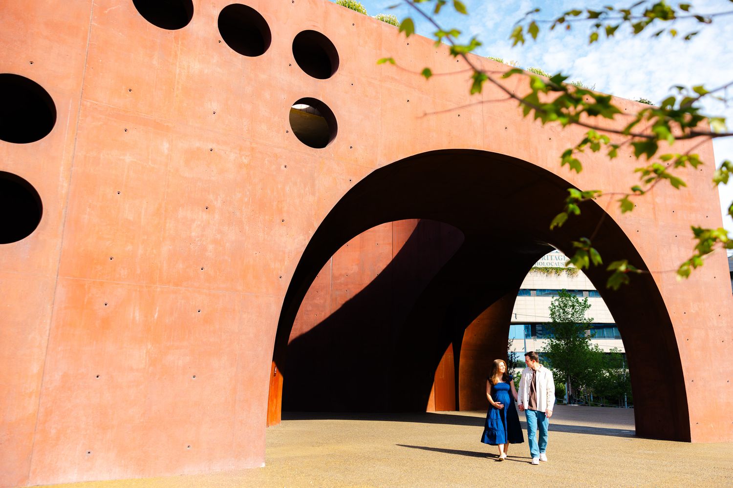 Couple walking under a large red sculptural arch with circular cutouts in an outdoor urban art installation.