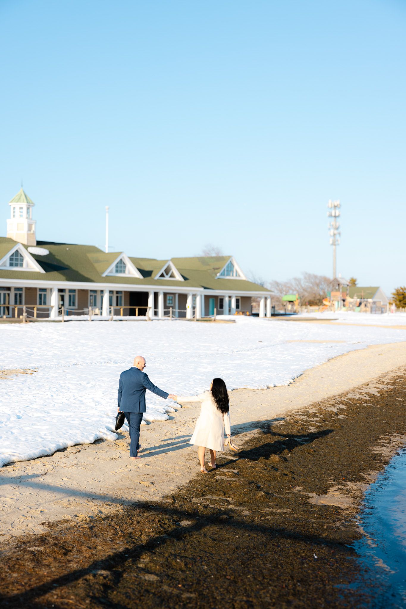 Couple holding hands walking along snowy winter beach with green roofed building in background on clear day.